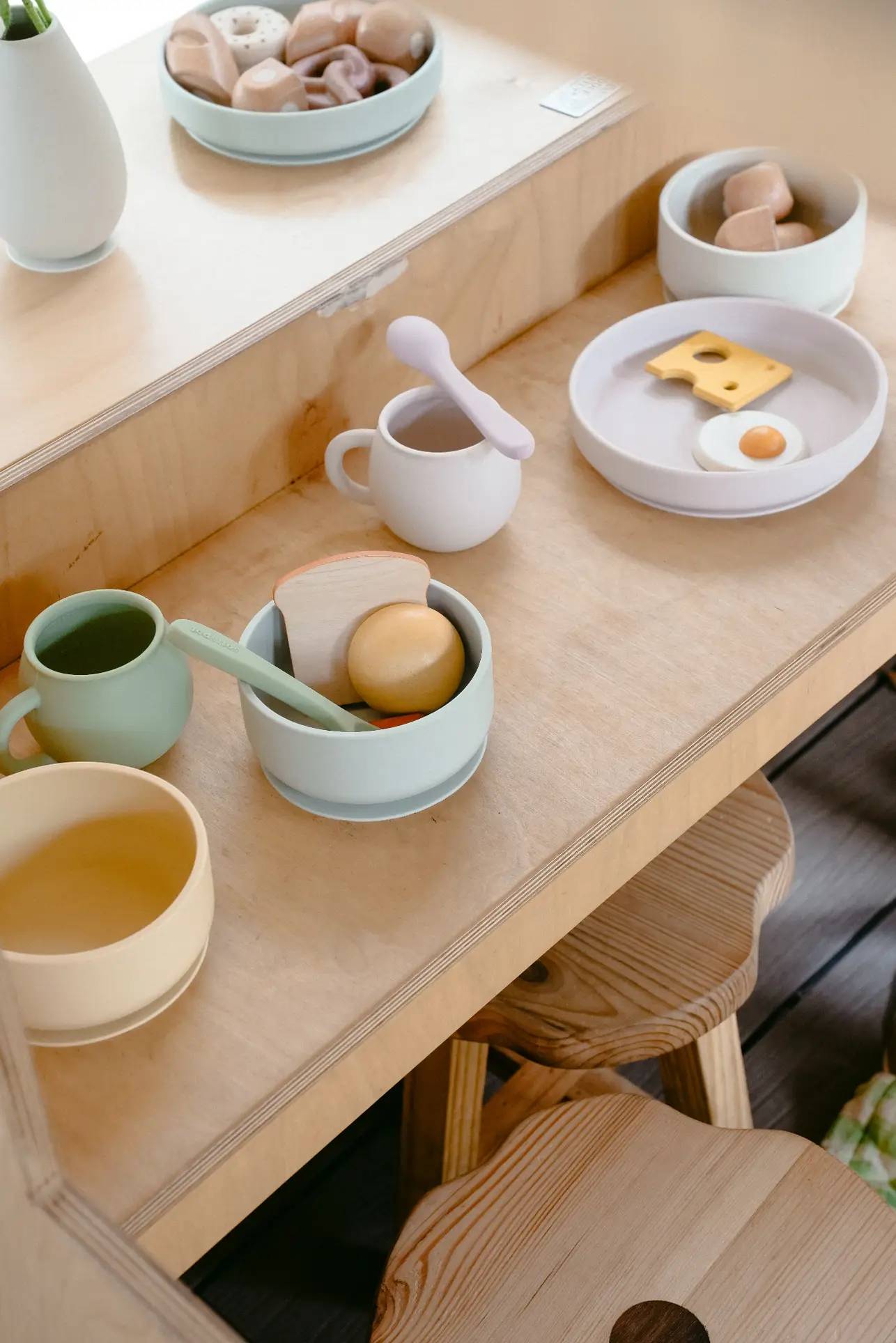 A children’s play kitchen is set with toy food and dishes, featuring the Soft Spot Soft Suction Bowl by Soft Spot made of food-grade silicone, along with plates of pretend eggs and cheese, cups, and utensils for a pretend tea party.
