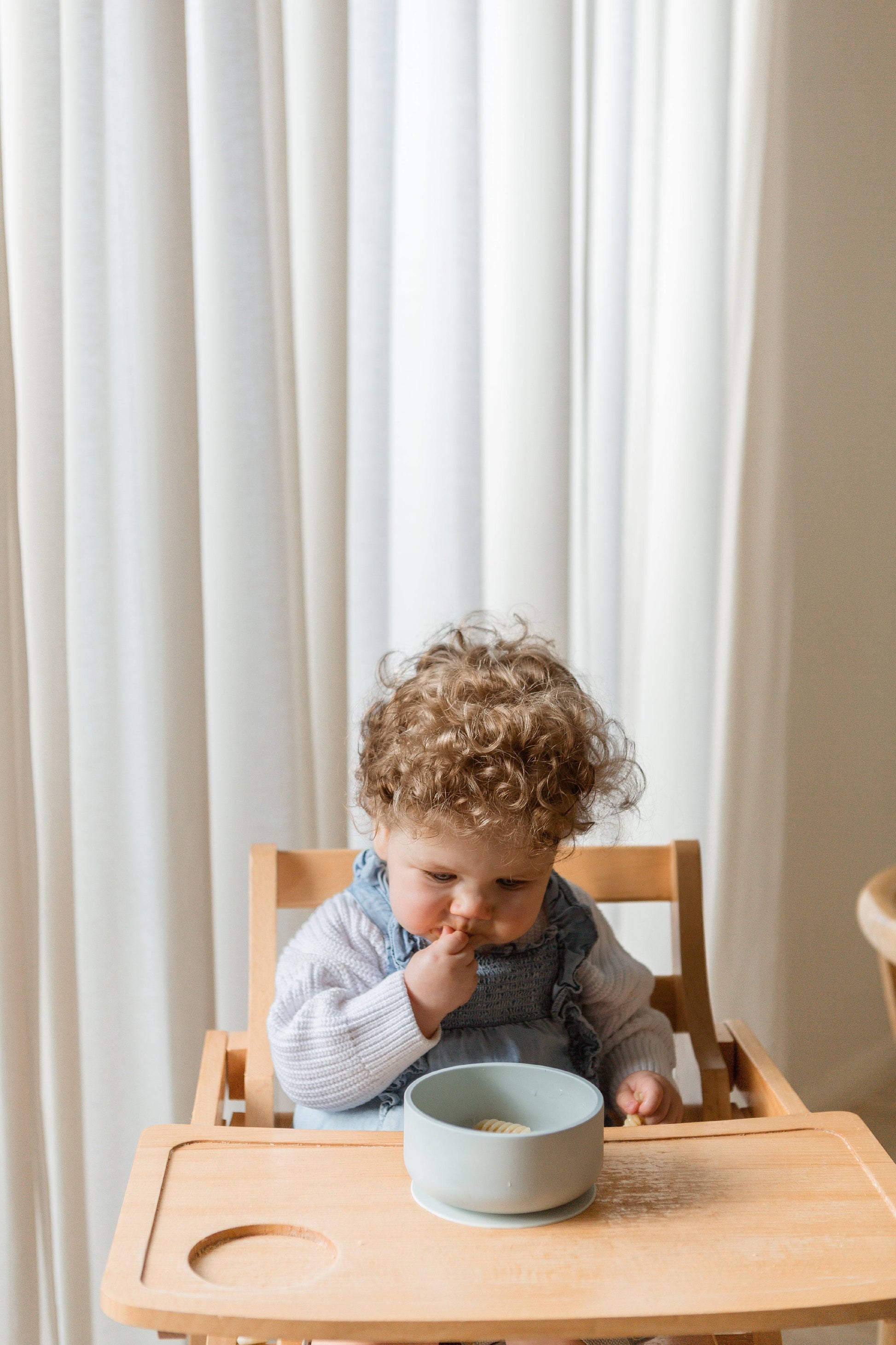 A curly-haired toddler in a light sweater and overalls enjoys a meal from the Soft Spot Soft Suction Bowl by Soft Spot, a light blue, food-grade silicone bowl with suction grip, while sitting in a wooden high chair with white curtains behind.