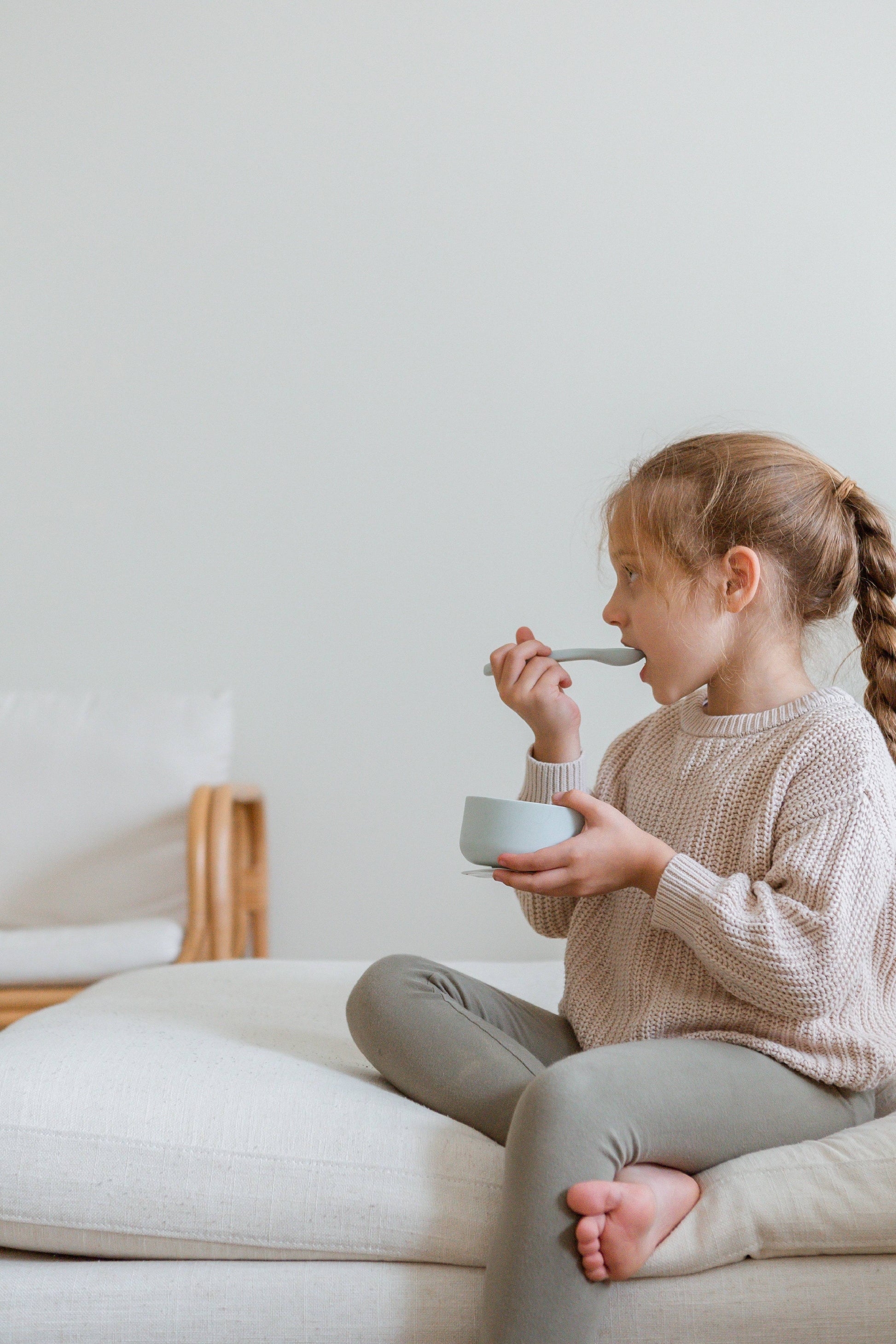 A young girl with a braided ponytail sits cross-legged on a light sofa, wearing a beige sweater and green leggings, eating from the Soft Spot Soft Suction Bowl by Soft Spot. The background is minimal and bright.