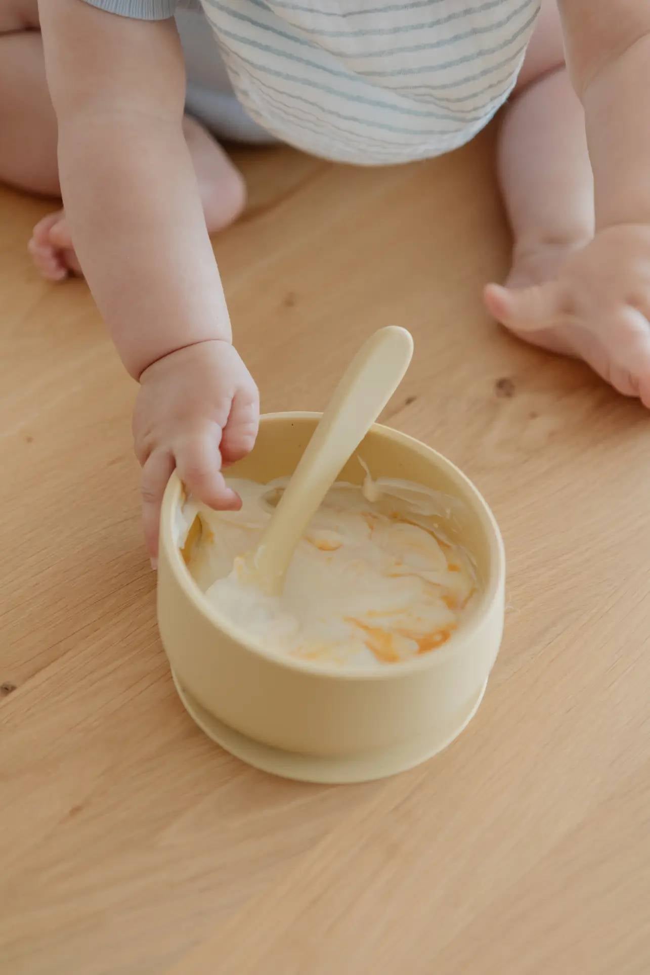 A baby’s arms, hands, and legs are shown as they reach for the Soft Spot Soft Suction Bowl by Soft Spot—a yellow food-grade silicone bowl with a matching spoon and strong suction grip—on a wooden floor. The bowl contains partially eaten food.