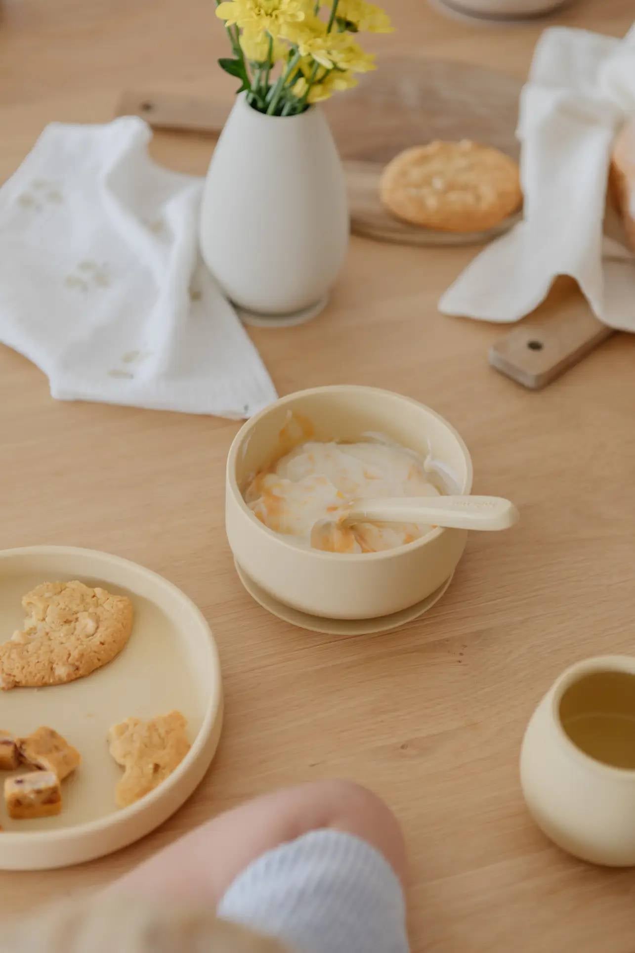 The Soft Spot Soft Suction Bowl by Soft Spot, in pale yellow food-grade silicone with a strong suction grip, holds yogurt and sauce. A matching spoon rests nearby on a wooden table with cookies, a cup, yellow flowers, napkin, and cutting board.