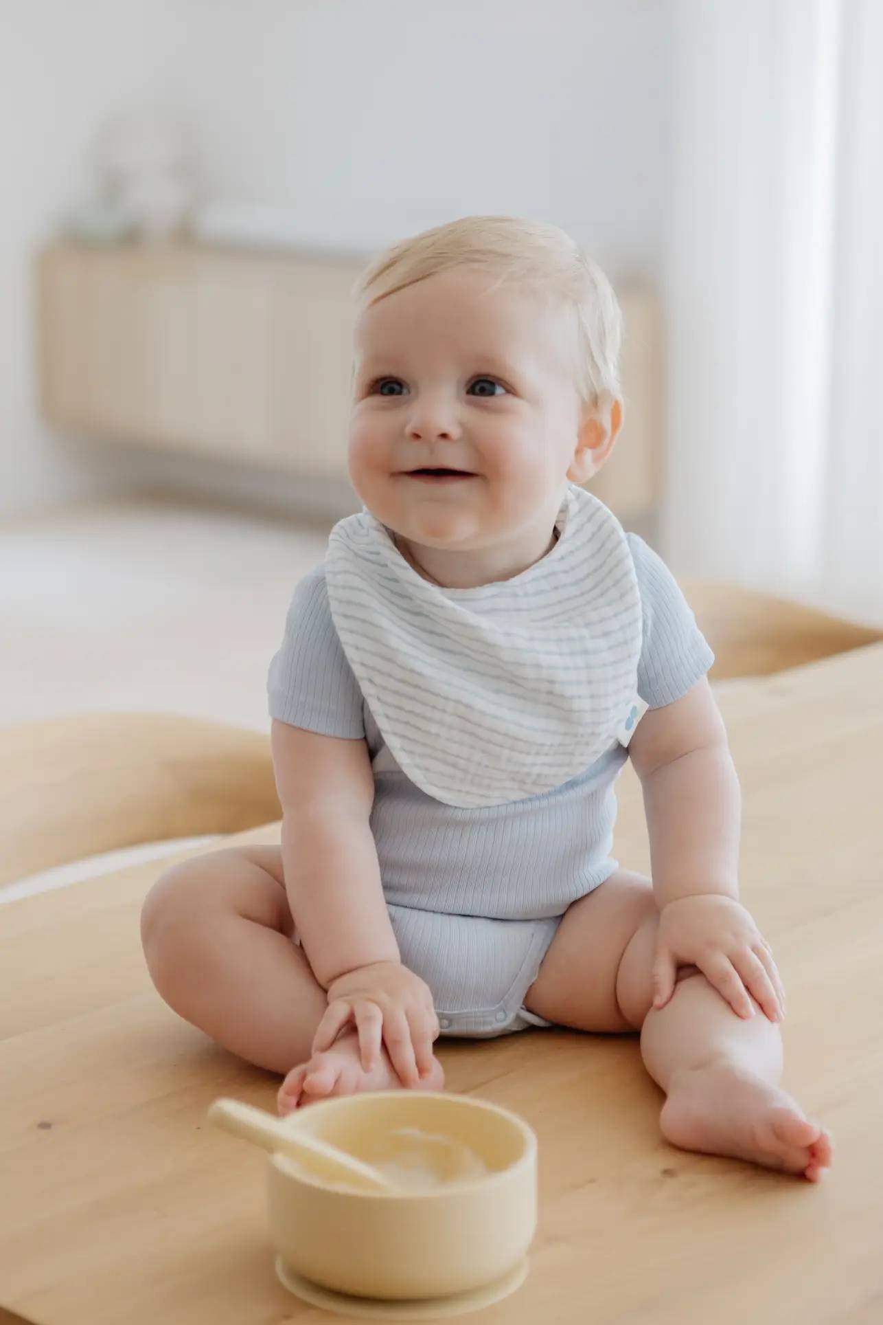 A blond baby smiles on a wooden table, wearing a light blue outfit and striped bib, with the Soft Spot Soft Suction Bowl and spoon from Soft Spot in front. The softly blurred background is bright.
