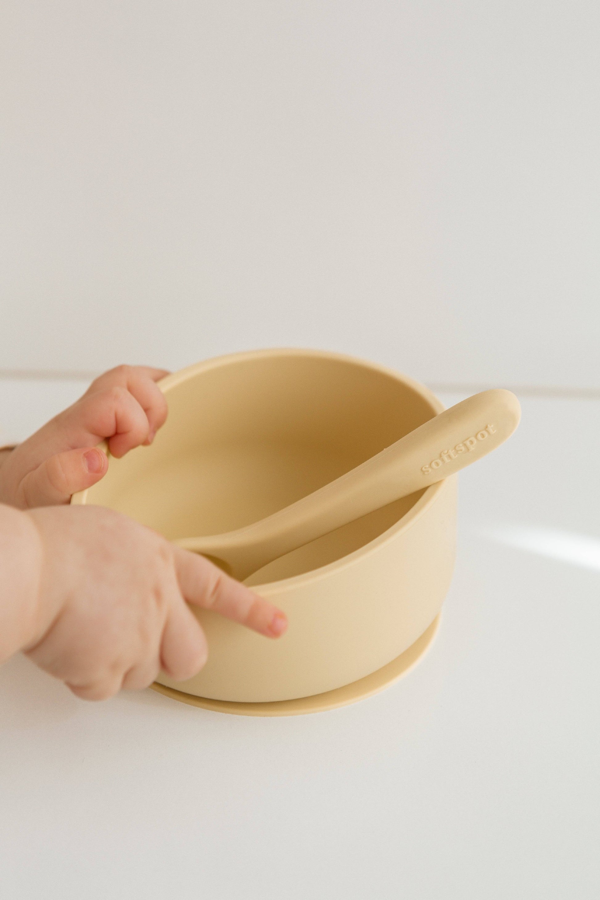 A child’s hands touch a beige Soft Spot Soft Suction Bowl with a matching spoon on a white surface. The minimalist design and secure suction grip from Soft Spot help prevent spills during mealtime.