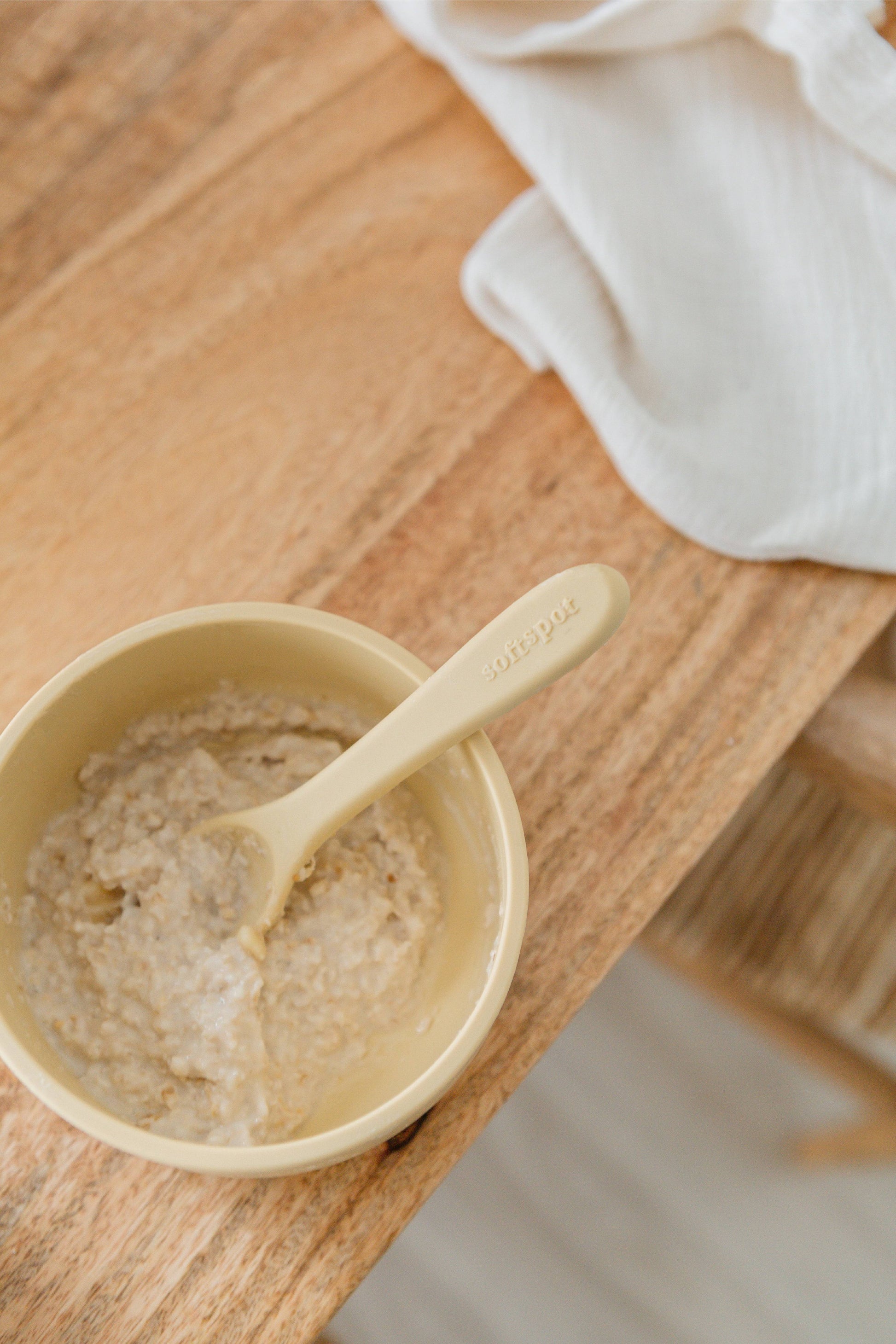 A beige Soft Spot Soft Suction Bowl filled with oatmeal and a matching spoon sits securely on a wooden table, its strong suction base holding it in place, with a white cloth nearby.