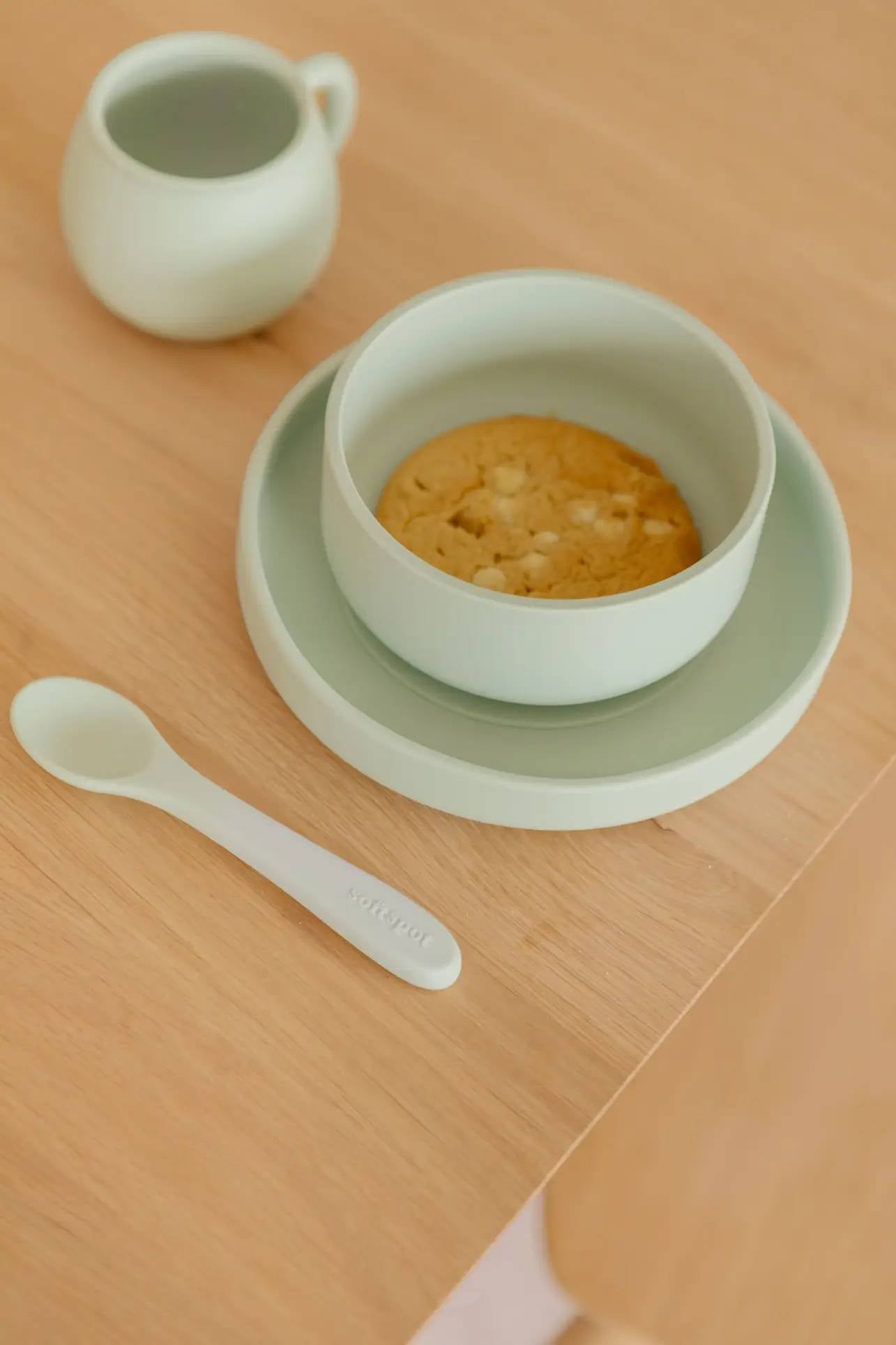A light green Soft Spot Soft Suction Bowl by Soft Spot, holding a cookie, along with a matching food-grade silicone cup, plate, and spoon are neatly arranged on a wooden table in soft natural light.
