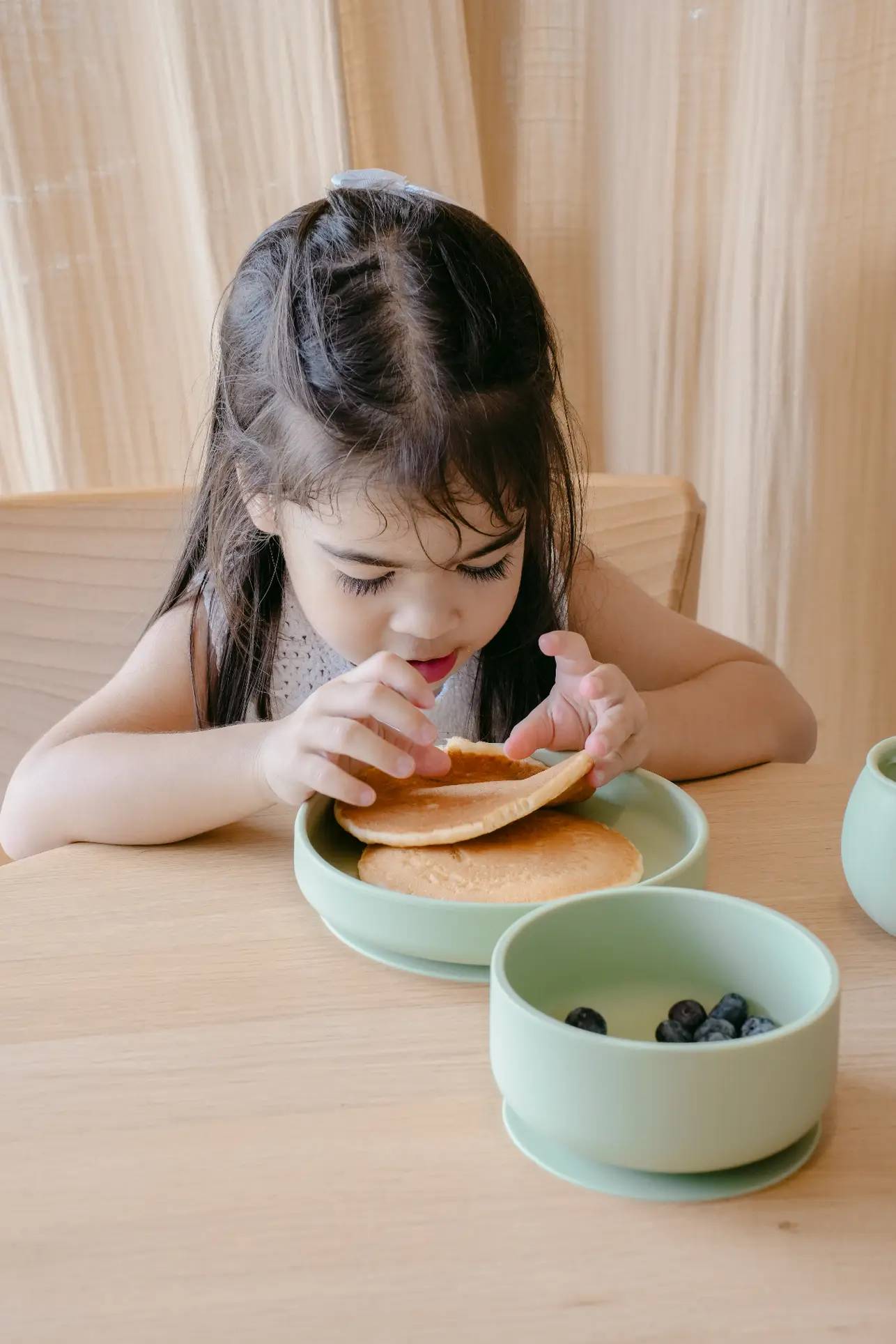 A young girl with long dark hair sits at a wooden table, holding pancakes on a plate. Next to her is a Soft Spot Soft Suction Bowl by Soft Spot, filled with blueberries. Light-colored curtains hang in the background.