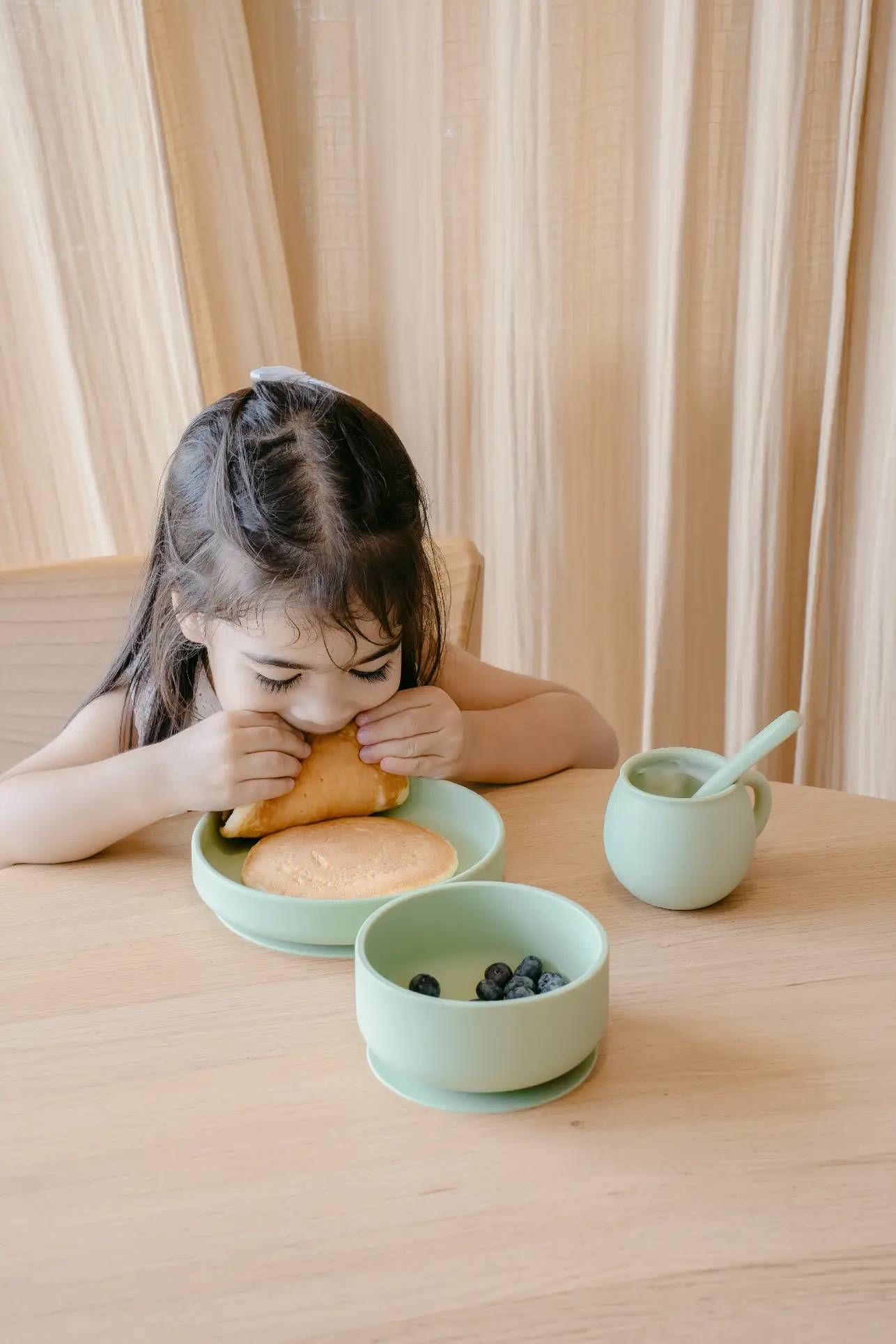 A young girl sits at a wooden table, eating a pancake from a green plate. In front of her are blueberries in a Soft Spot Soft Suction Bowl by Soft Spot, and a green cup with a spoon. Beige curtains hang in the background.