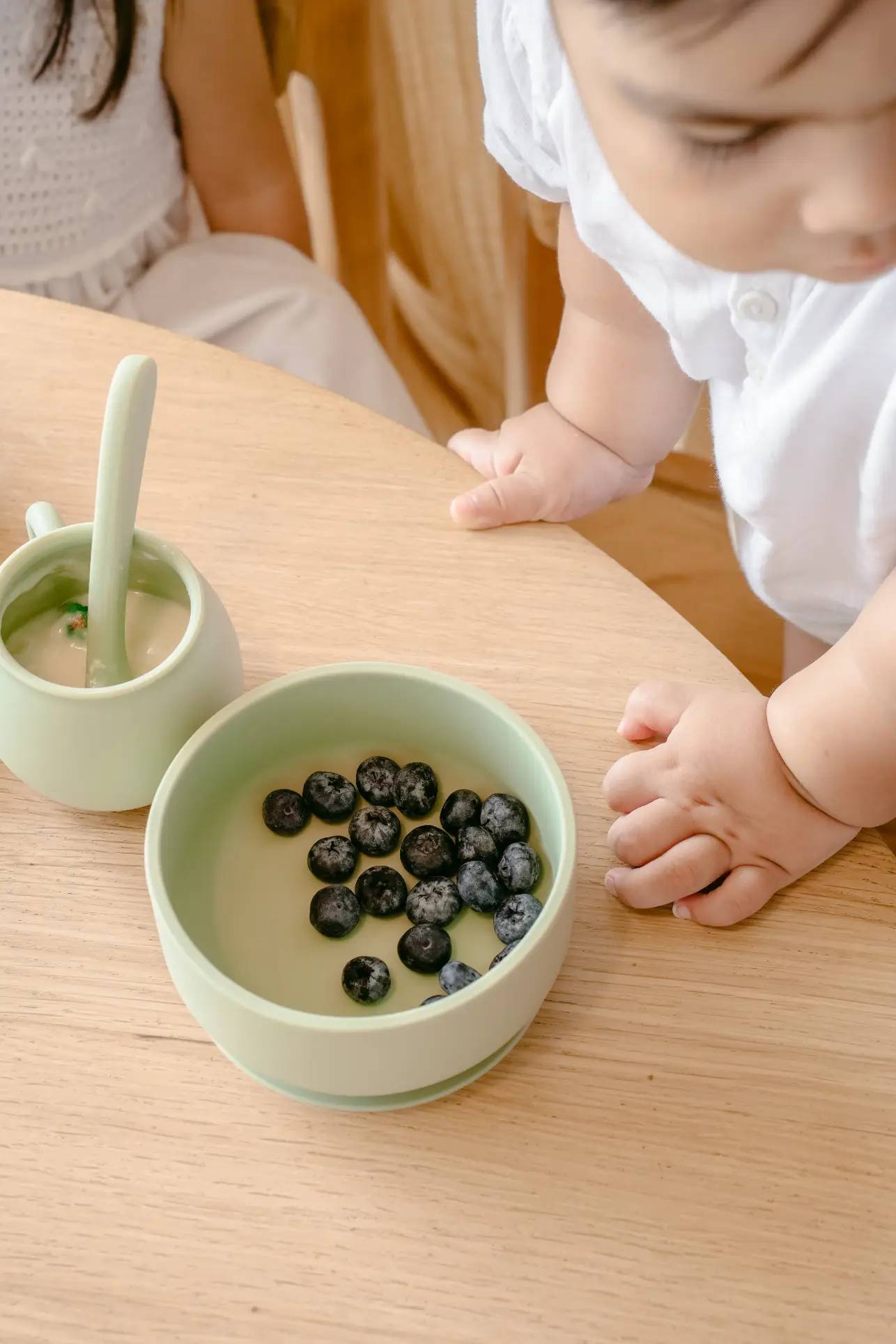 A young child in a white shirt leans over a wooden table with a Soft Spot Soft Suction Bowl by Soft Spot filled with blueberries and a matching cup and spoon. Another child sits nearby, partially visible.