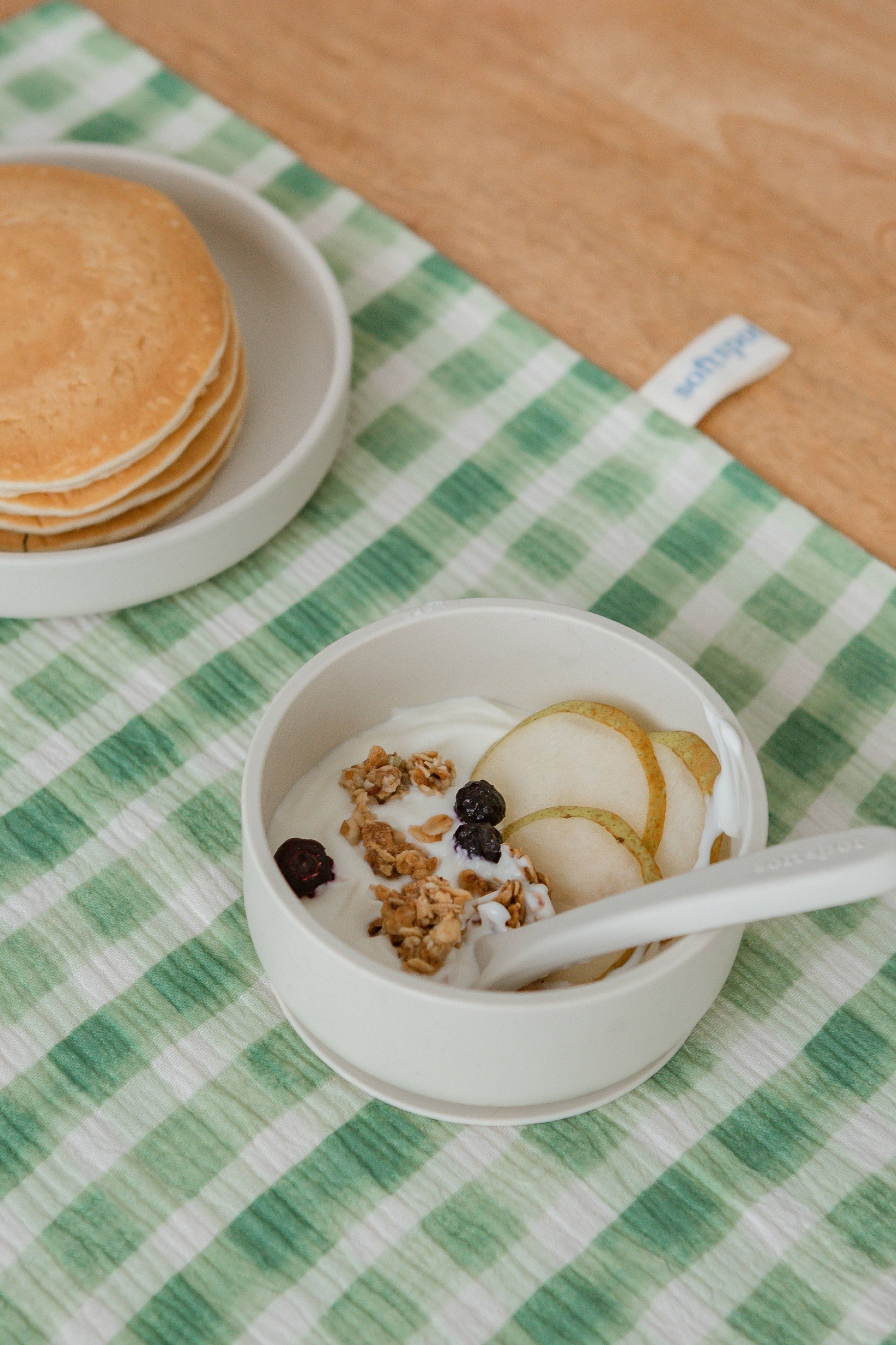 A Soft Spot Soft Suction Bowl by Soft Spot, filled with yogurt, granola, pear slices, and dried berries, sits on a green plaid cloth next to stacked pancakes. A spoon rests in the bowl, steadied by its suction base.