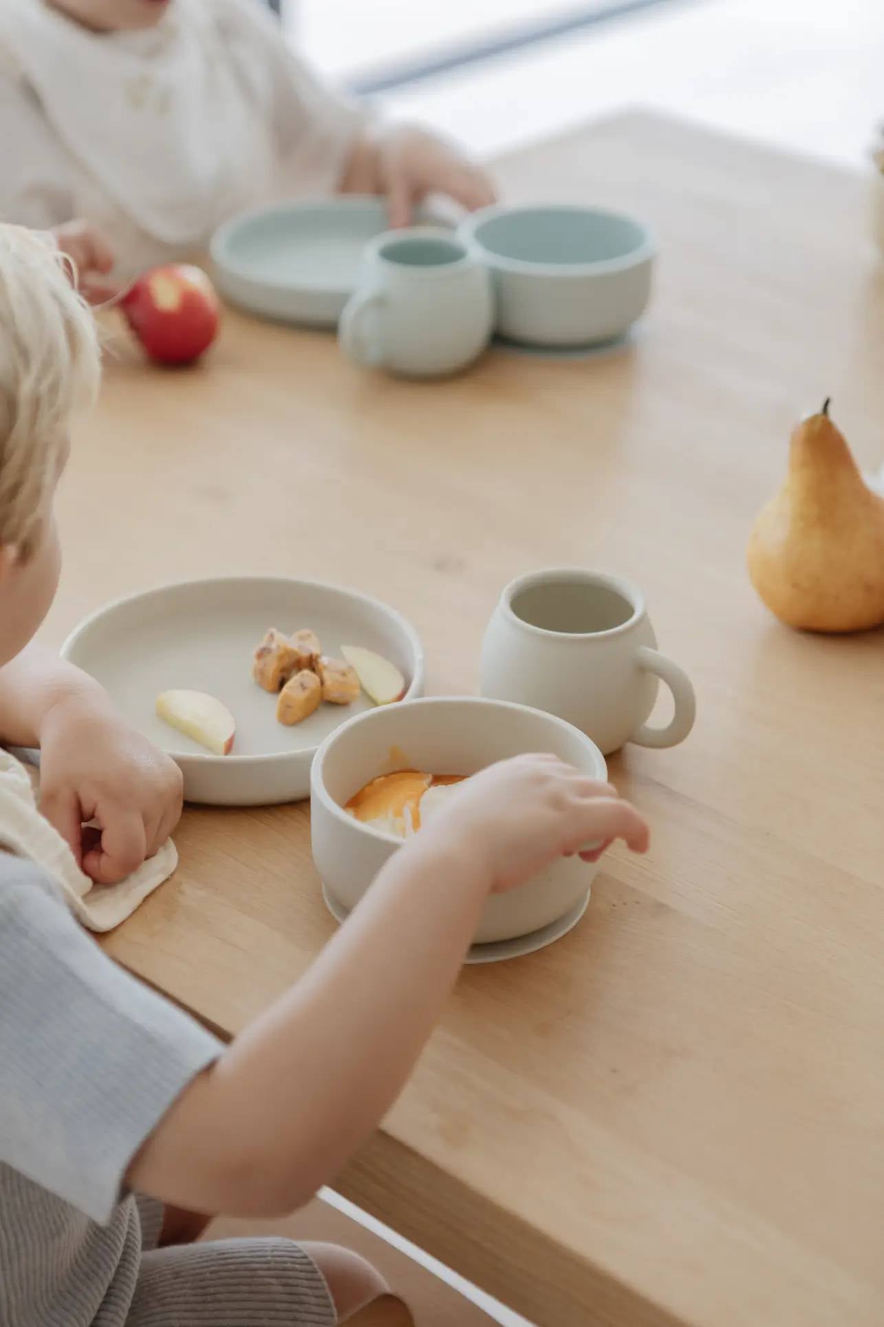 Two young children sit at a wooden table using Soft Spot Soft Suction Bowls by Soft Spot, made from food-grade silicone. One child enjoys snacks like apple slices and crackers in this calm, minimalistic setting.