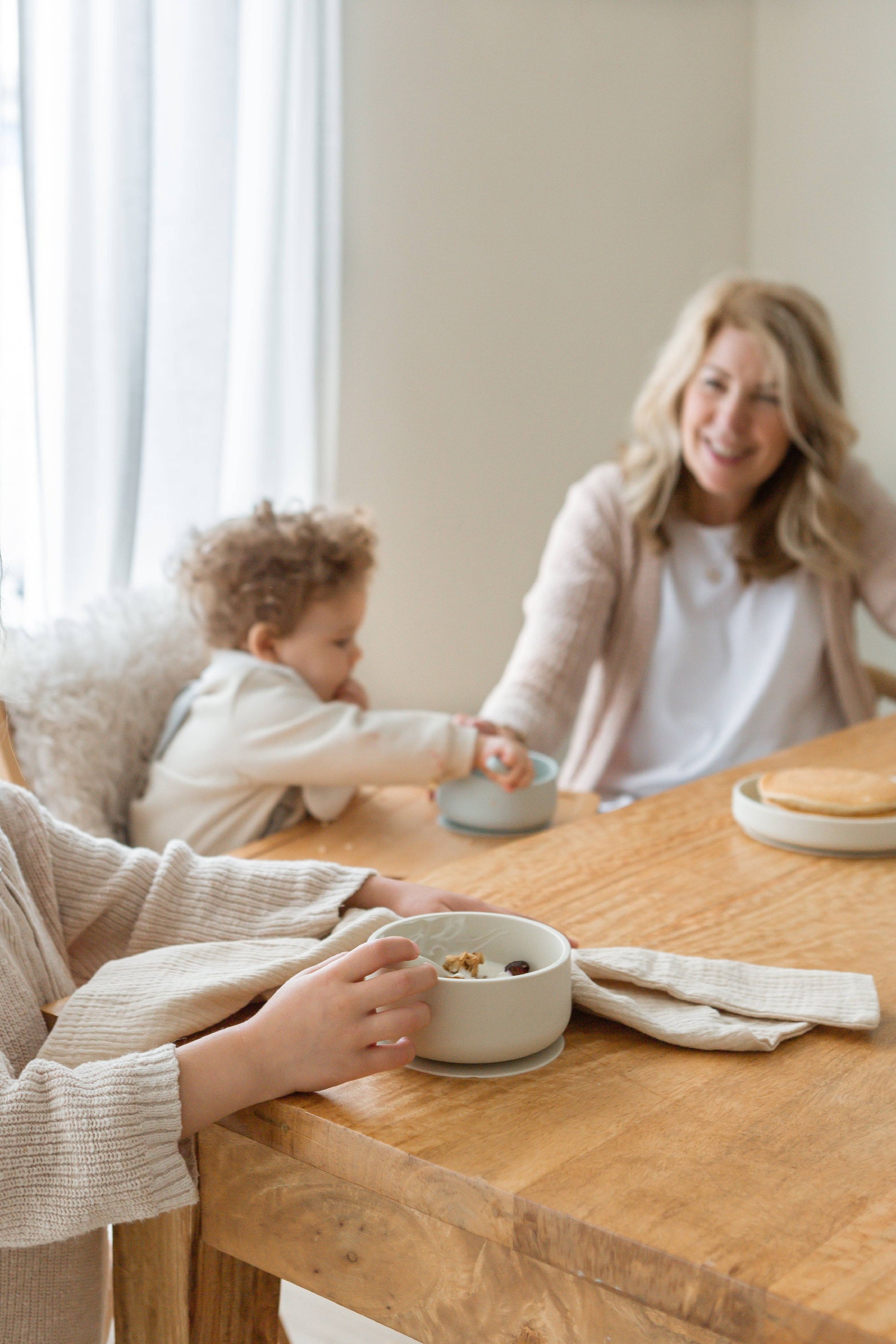 A woman and young child smile at a wooden table with Soft Spot Soft Suction Bowls, reaching for each other in a cozy dining area filled with delicious pancakes.