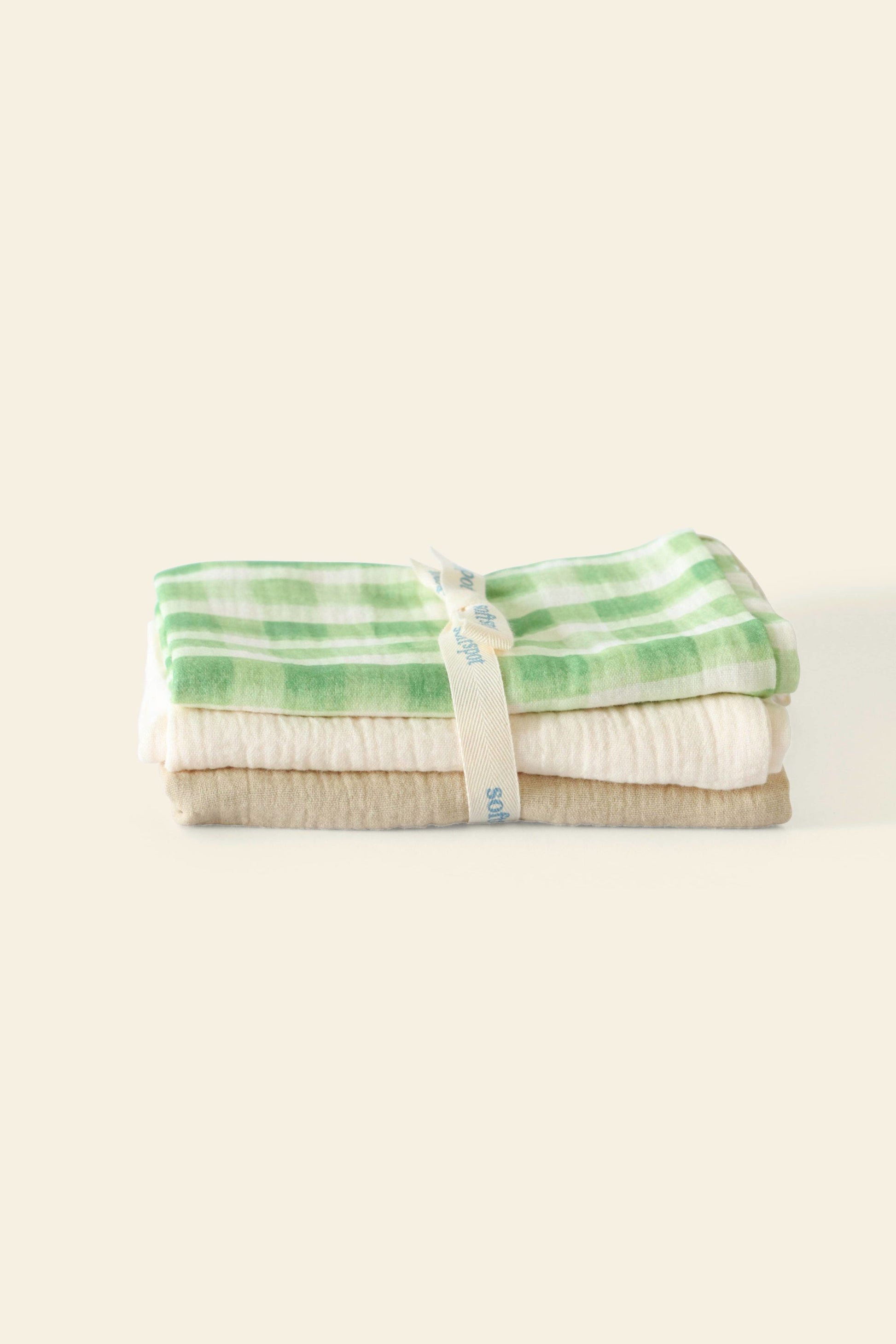 The Soft Spot Soft Tea Towels (Set of 3) by Soft Spot features a neatly folded trio tied with a cream ribbon; the top towel displays a green and white checkered pattern, while the others are solid neutrals, set against a plain background.