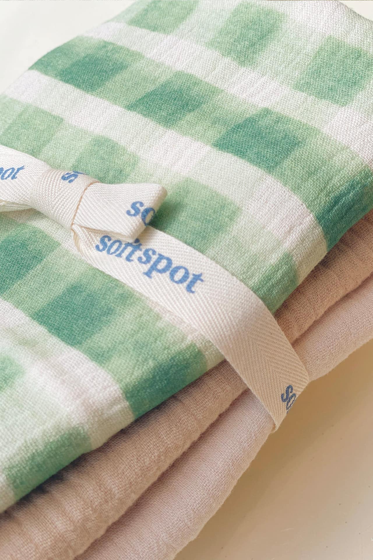A close-up of Soft Spots Soft Tea Towels (Set of 3): the top towel features a green and white check pattern, tied with a cream ribbon with blue spots. A beige towel from the set, ideal as a placemat or hand towel, peeks out underneath.