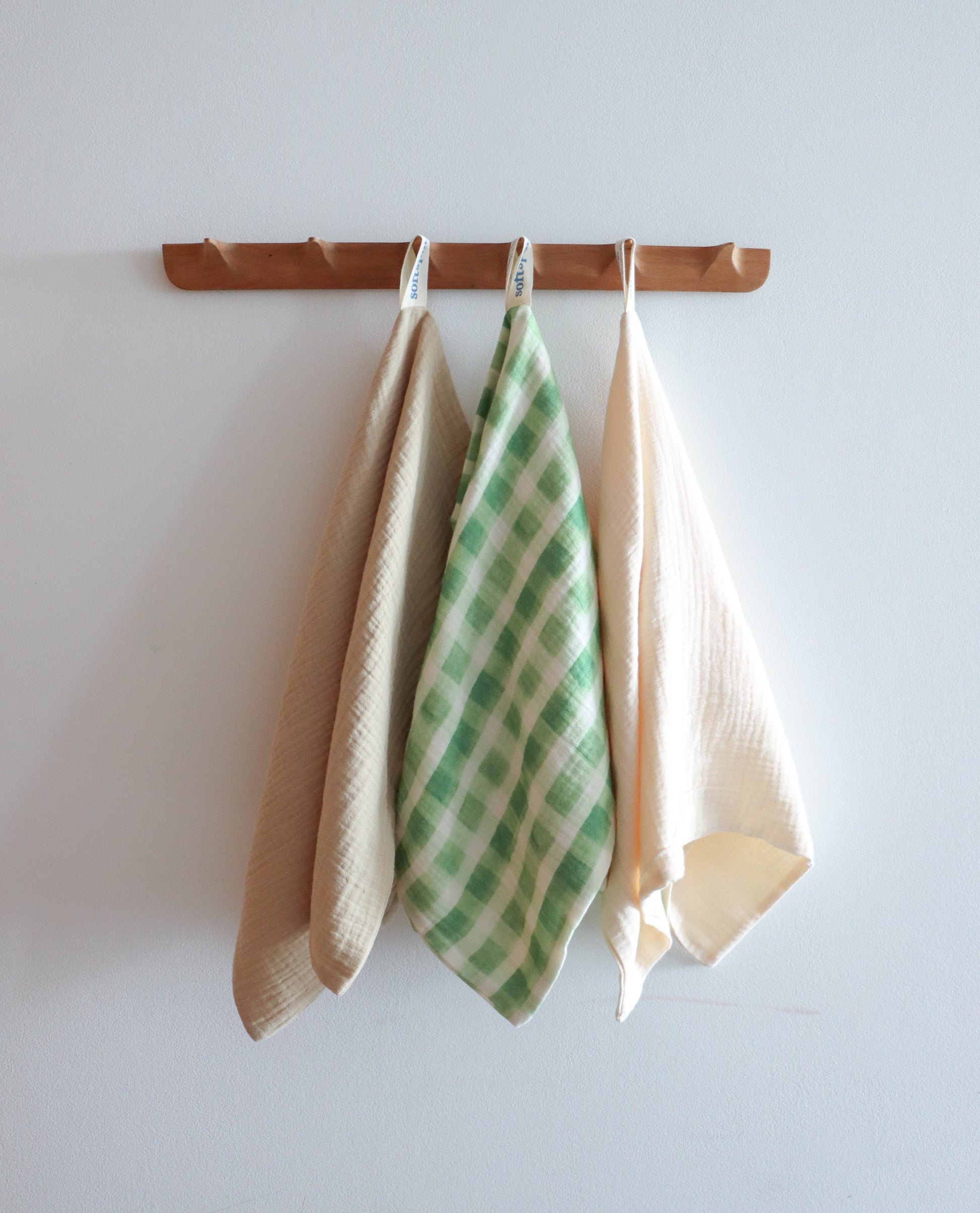The Soft Spot Soft Tea Towels (Set of 3) by Soft Spot—featuring a soft tea towel, a green and white checkered hand towel, and an off-white towel—hang on a wooden rack against a plain white wall.