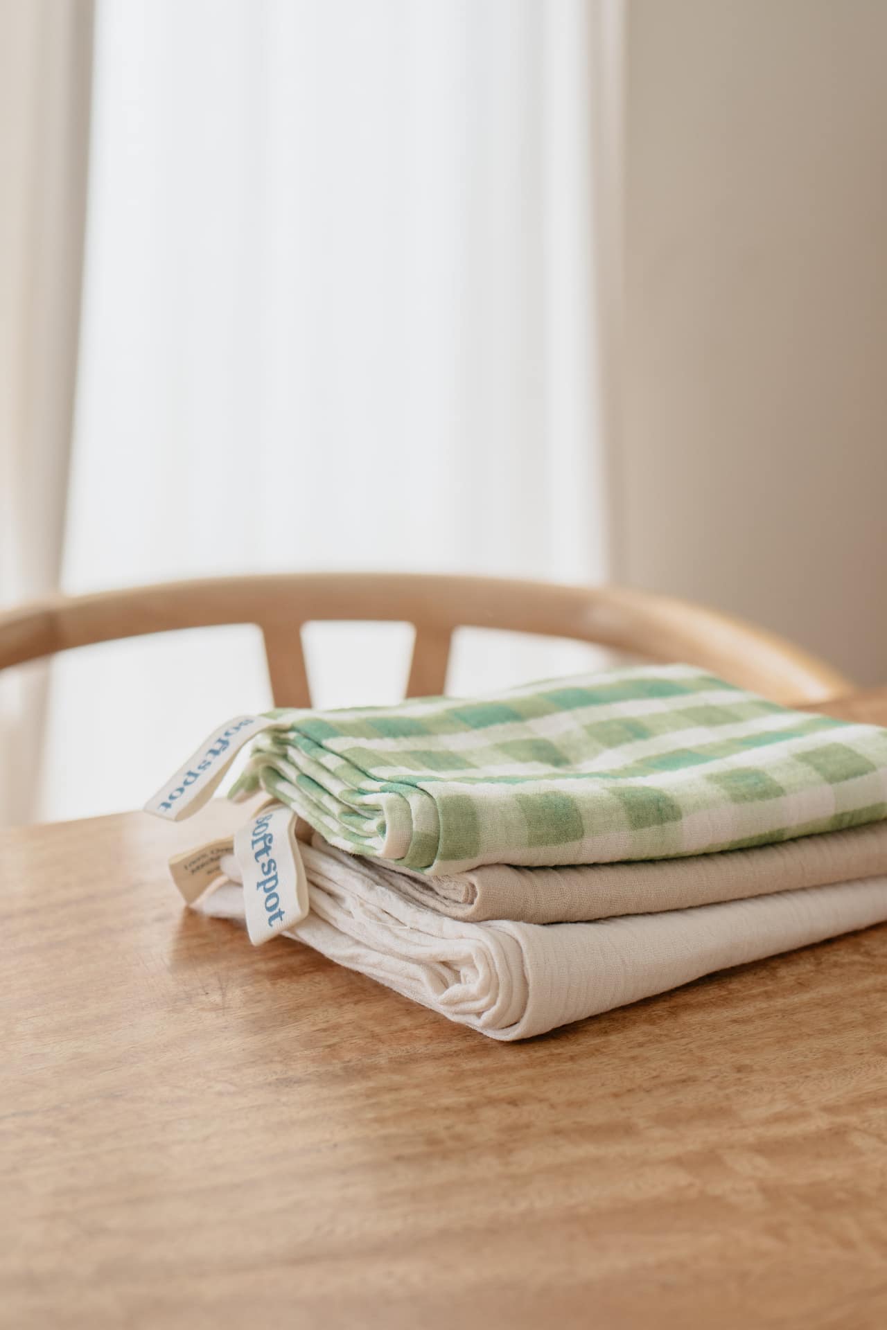 Three neatly folded Soft Spot Soft Tea Towels from Soft Spot, including a green and white checkered design and a solid white one, are stacked on a wooden table with a chair and sheer white curtains in the background.
