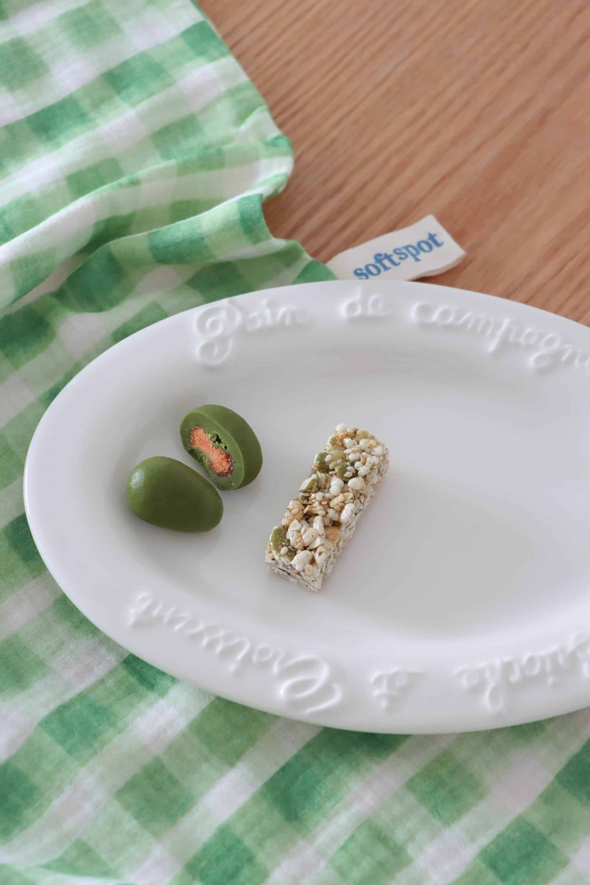 A white plate with two green chocolate-covered almonds, one halved, and a nut and seed bar sits atop a Soft Spot Soft Tea Towel (Set of 3) by Soft Spot, arranged on a wooden surface.