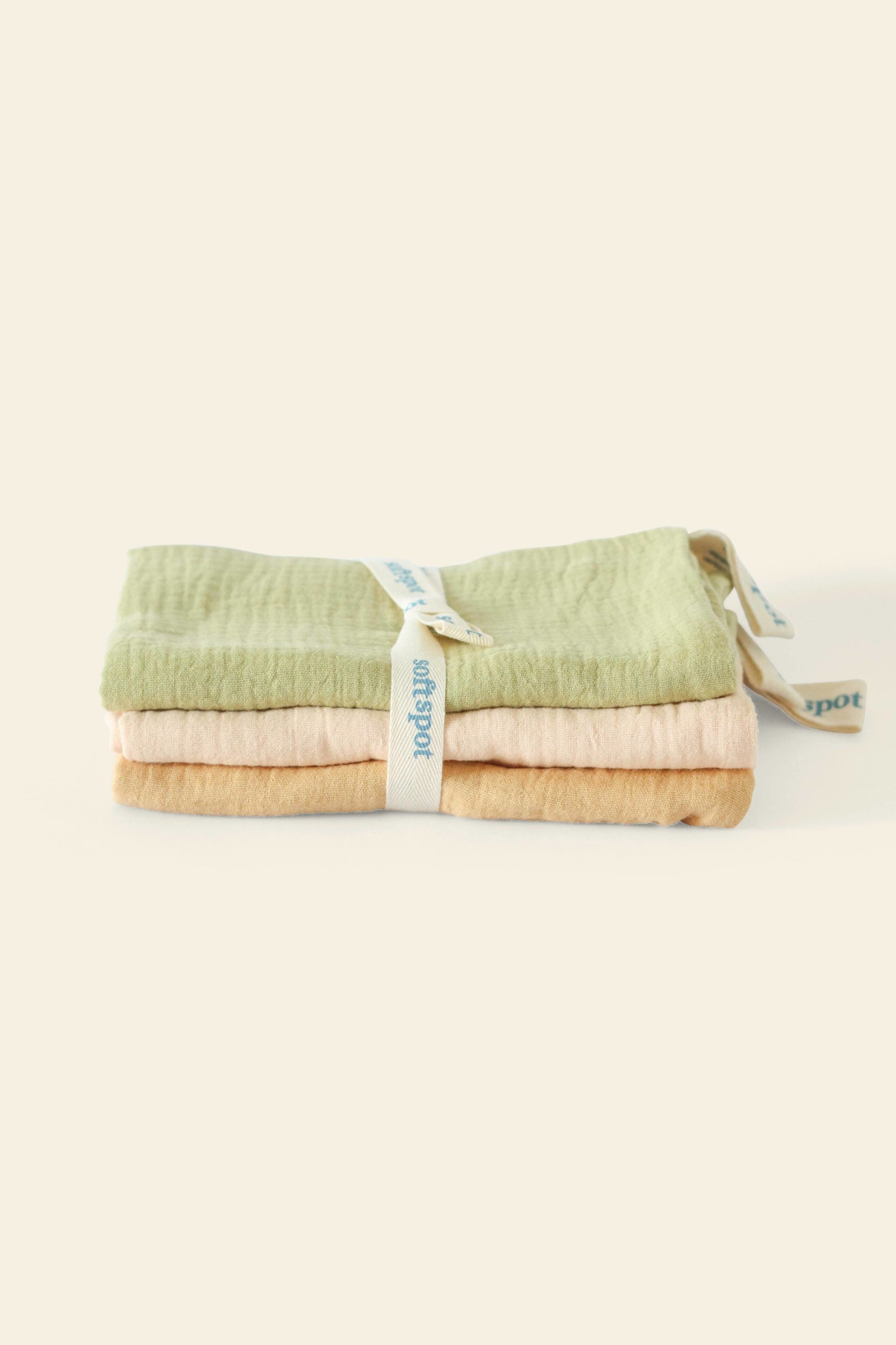The Soft Spot Soft Tea Towels (Set of 3) by Soft Spot features folded towels in pastel green, peach, and beige stacked together and secured with a white branded ribbon, ideal for use as hand or tea towels.