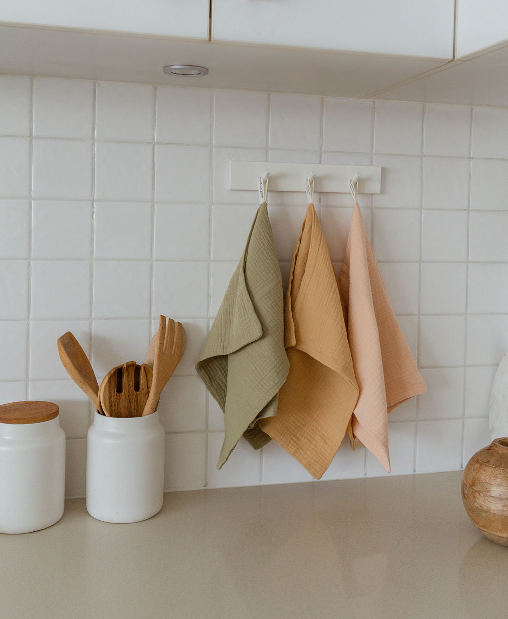 Three Soft Spot Soft Tea Towels (Set of 3) by Soft Spot hang on white hooks against a tiled kitchen wall, near two white jars—one with wooden utensils—on a beige countertop.
