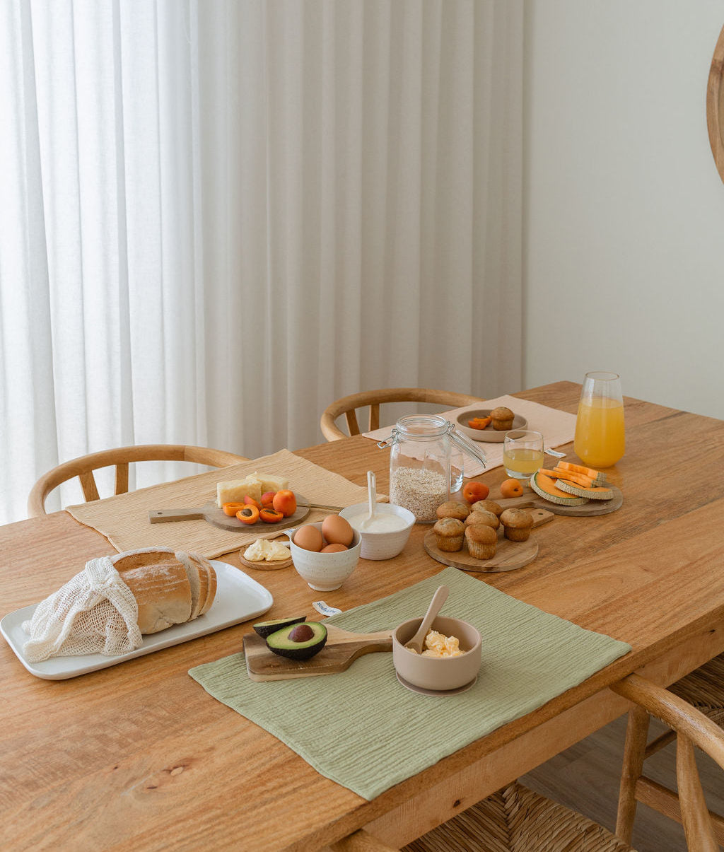 A wooden dining table set for breakfast features Soft Spot Soft Tea Towels (Set of 3) by Soft Spot among bread, butter, eggs, avocado, fruit, a jar of oats, and a glass of orange juice. Wooden chairs and soft light complete the inviting scene.