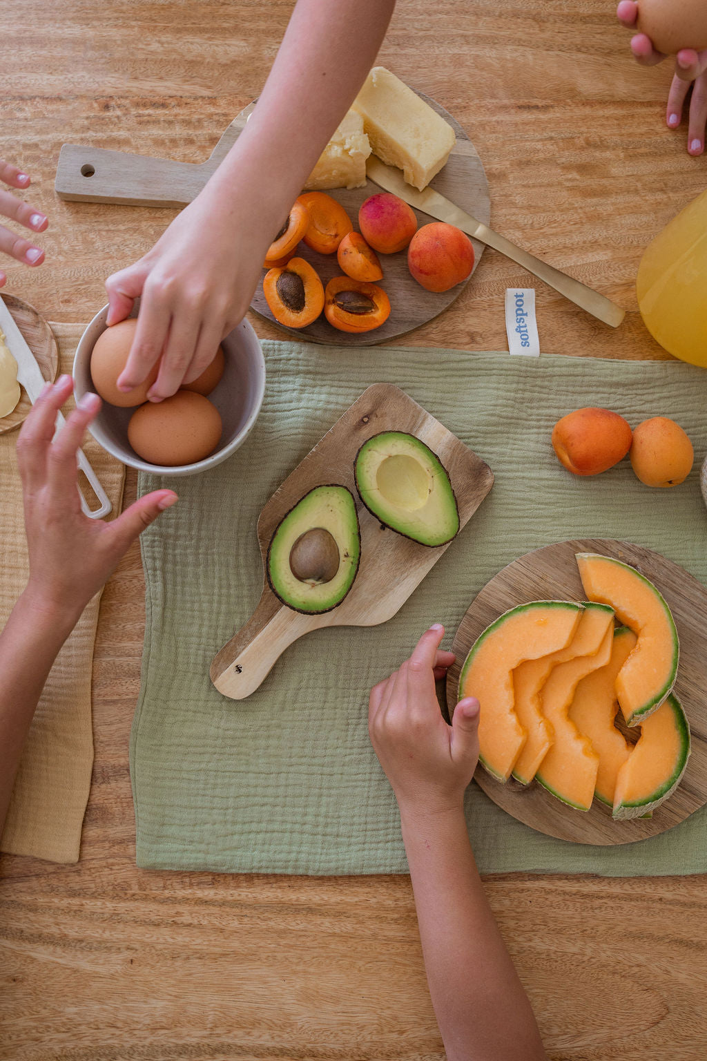 Top-down view of hands reaching for eggs, avocado, apricots, cheese, and cantaloupe on a wooden table with cutting boards, a green placemat, and a neatly folded Soft Spot Soft Tea Towel from the Soft Spot Set of 3.