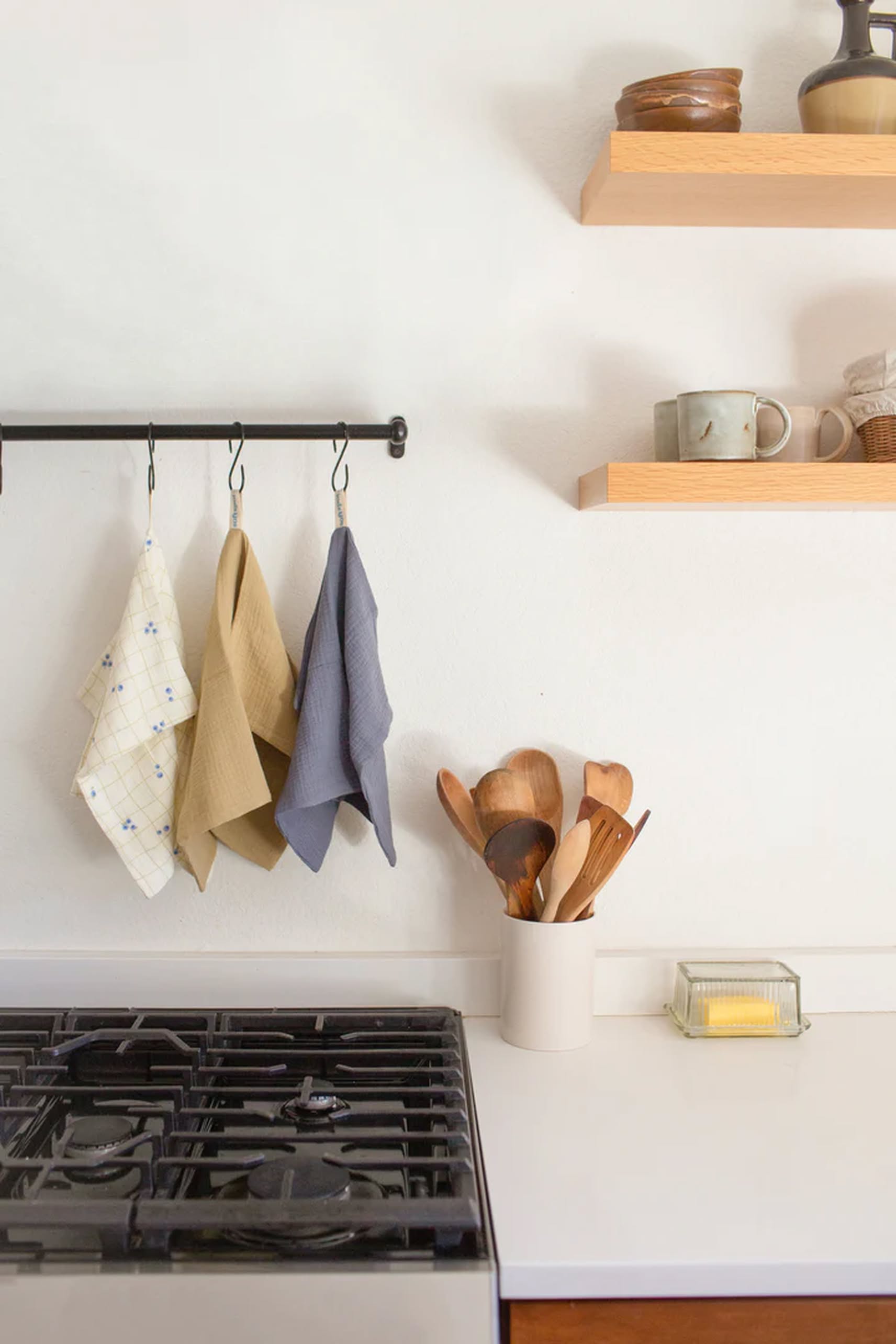 A kitchen countertop with a stove, three Soft Spot Soft Tea Towels by Soft Spot hanging from hooks, a white container of wooden utensils, a butter dish, and two wooden shelves with bowls, mugs, and small items on a white wall.
