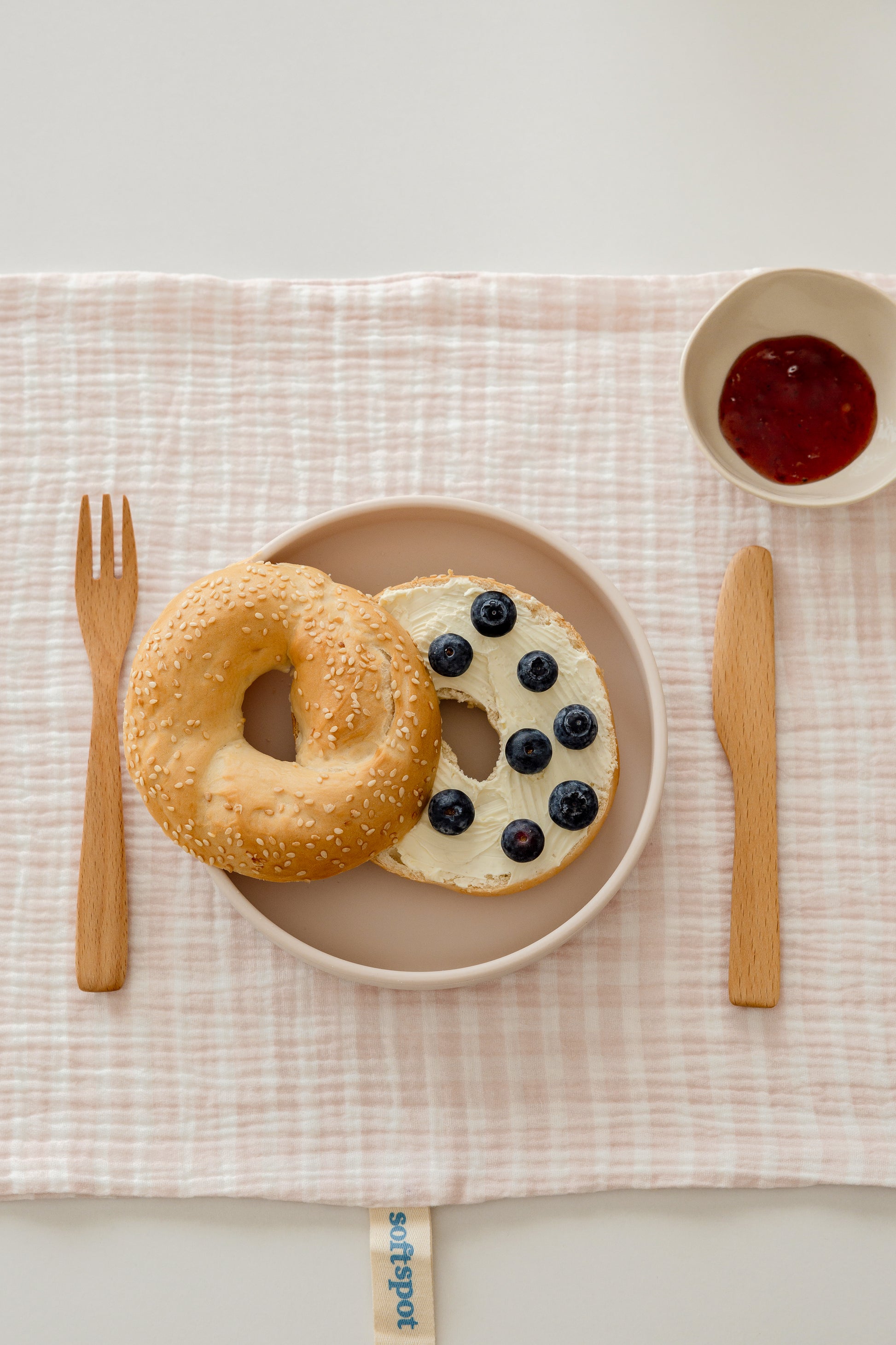 A sesame bagel with cream cheese and blueberries sits on a Soft Spot Soft Tea Towel by Soft Spot, alongside a wooden knife, fork, and a bowl of red jam on the soft, textured fabric.