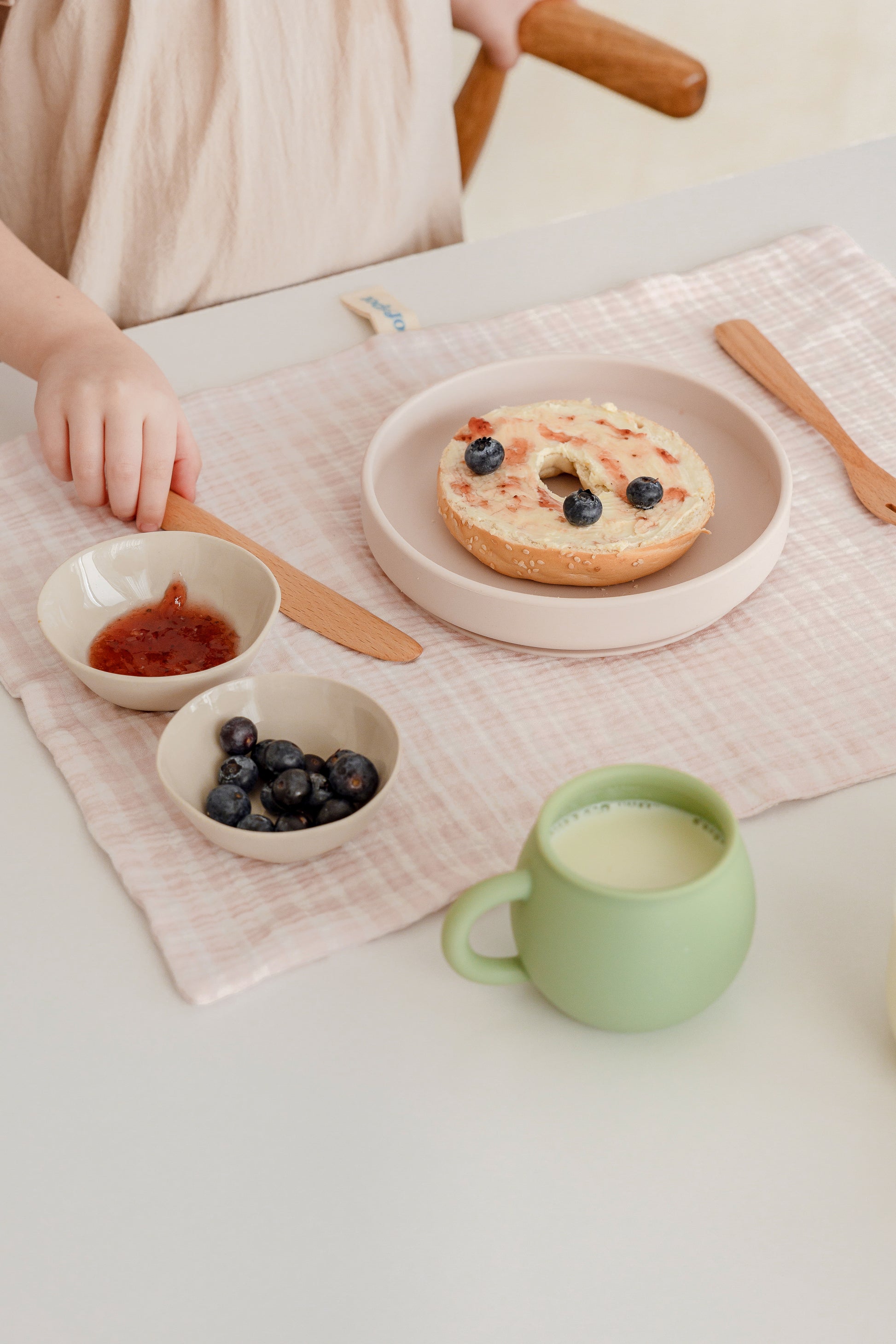 A child sits at a table set with a bagel topped with blueberries, bowls of jam and berries, a wooden knife, green cup of milk, and a vibrant pink Soft Spot Soft Tea Towel (Set of 3) placemat by Soft Spot brightening the breakfast scene.