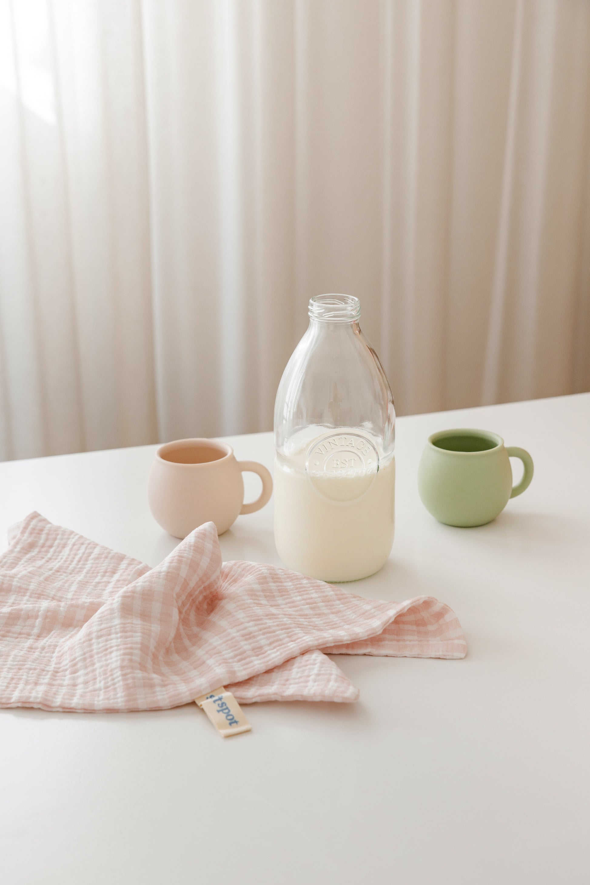 A glass bottle of milk sits on a white table beside Soft Spot Soft Tea Towels (Set of 3) and two small ceramic cups, one pink and one green. Light curtains in the background add a soft, airy touch.