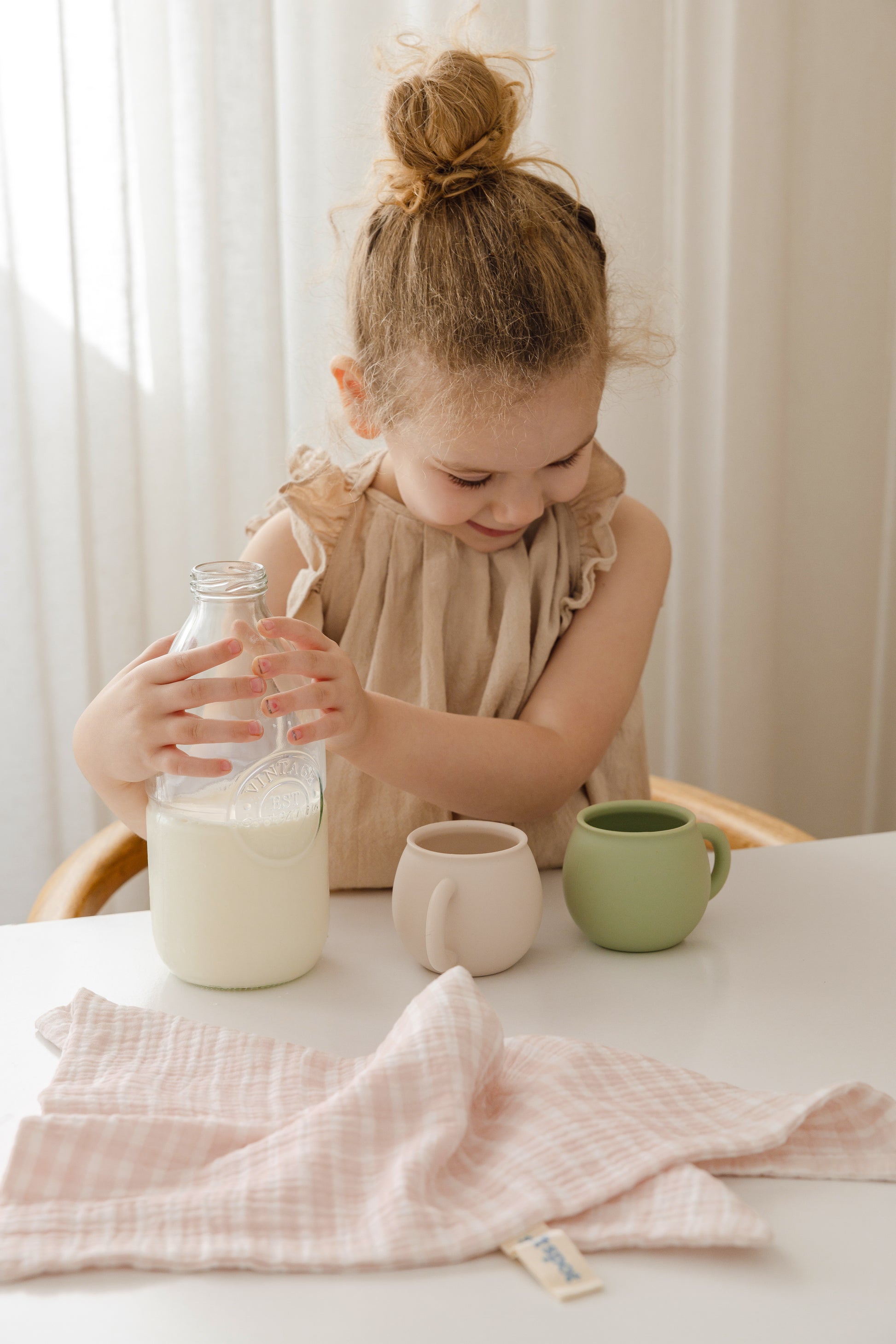 A young girl with light brown hair in a bun smiles while pouring milk into a mug at a white table, with a green cup and Soft Spot Soft Tea Towels (Set of 3) by Soft Spot nearby.