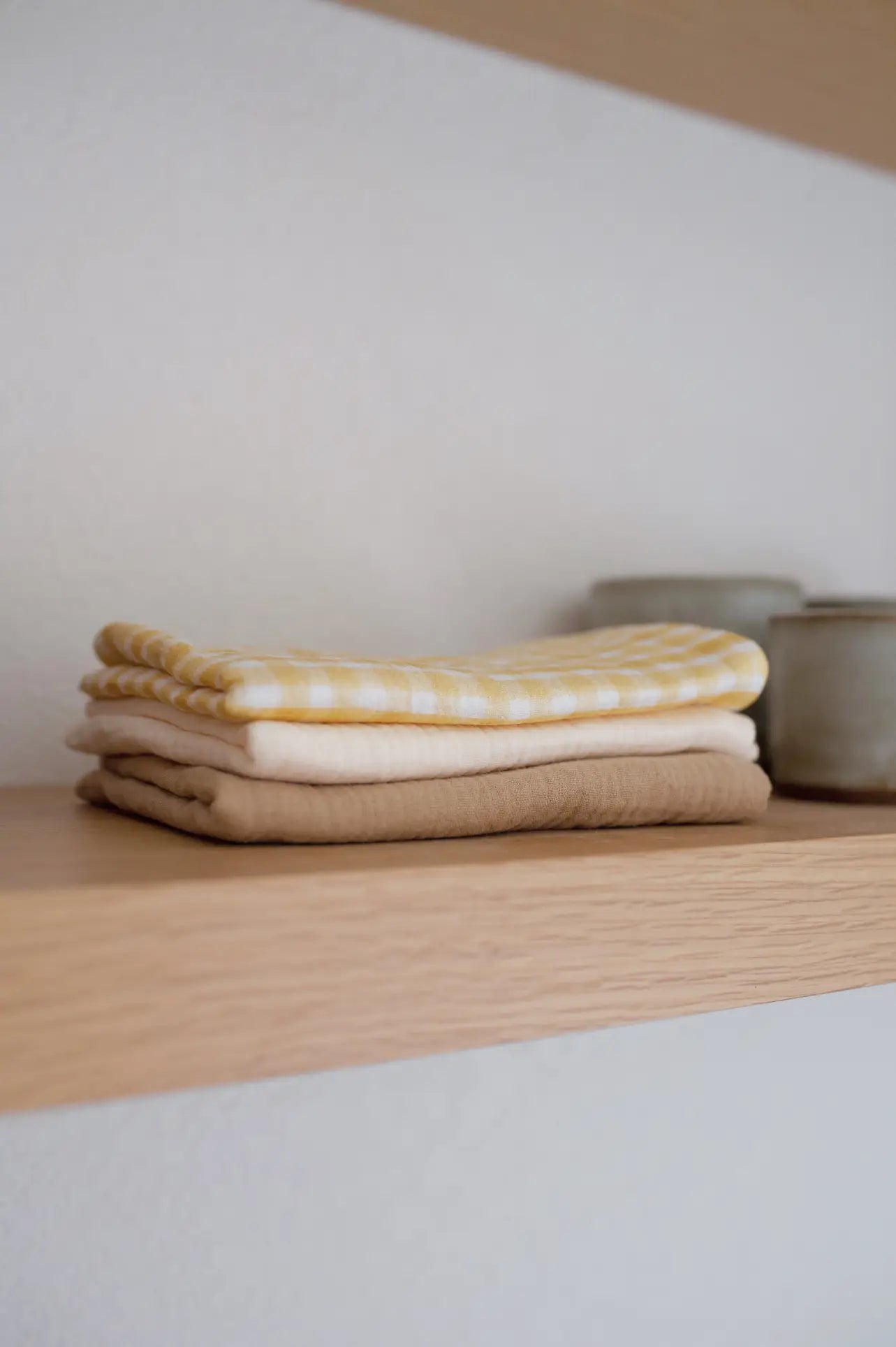 A stack of Soft Spot Soft Tea Towels (Set of 3) in beige, white, and yellow patterns rests on a wooden shelf beside two small ceramic containers.