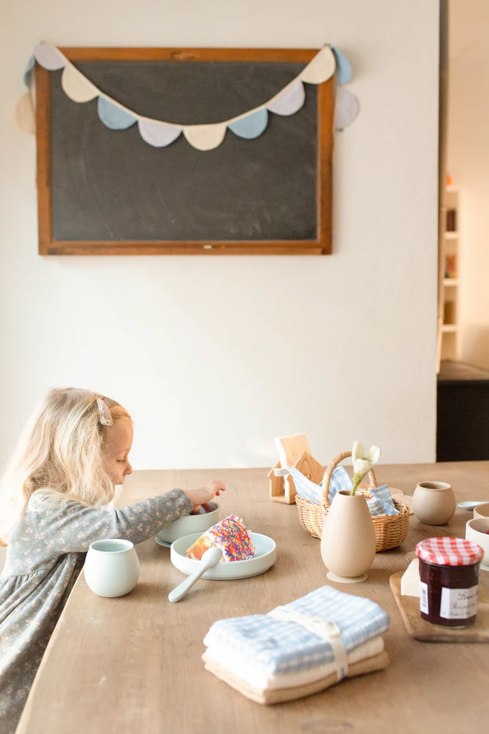 A young girl eats at a wooden table decorated with Soft Spot Soft Tea Towels, jars, napkins, and a flower vase. Behind her, pastel bunting hangs above a chalkboard as warm light fills the cozy room.