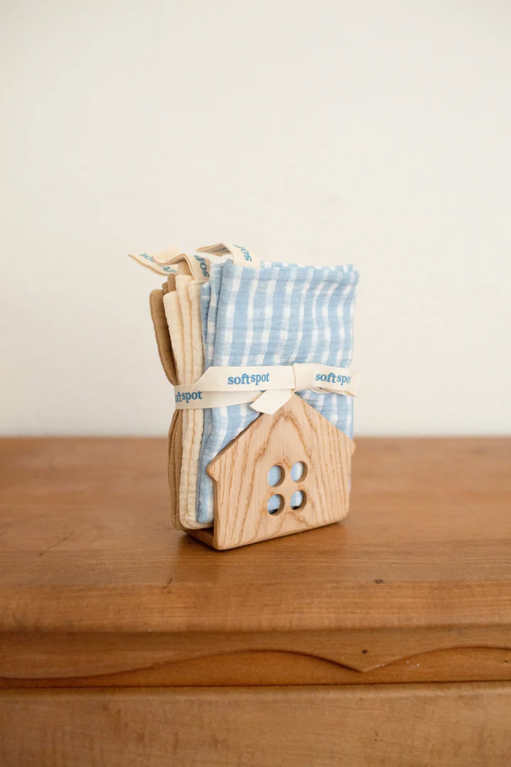 The Soft Spot Soft Tea Towels (Set of 3) by Soft Spot, folded and tied with a ribbon, are displayed upright in a wooden house-shaped holder on a wooden surface against a plain light background.