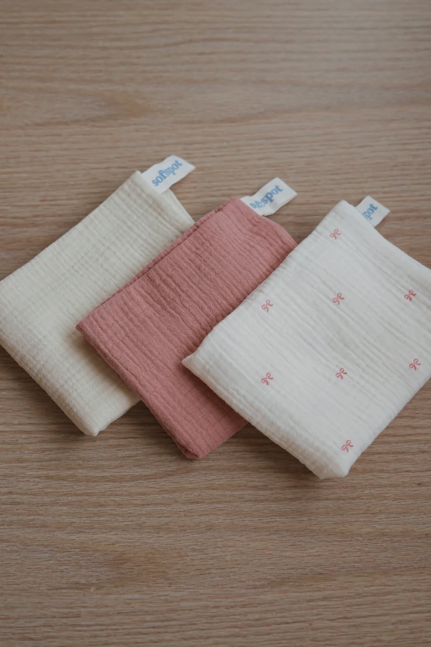 The Soft Spot Soft Tea Towels (Set of 3) by Soft Spot, in cream, pink, and cream with pink bows—each featuring a white tag—are neatly folded as hand, face, or tea towels on a wooden surface.
