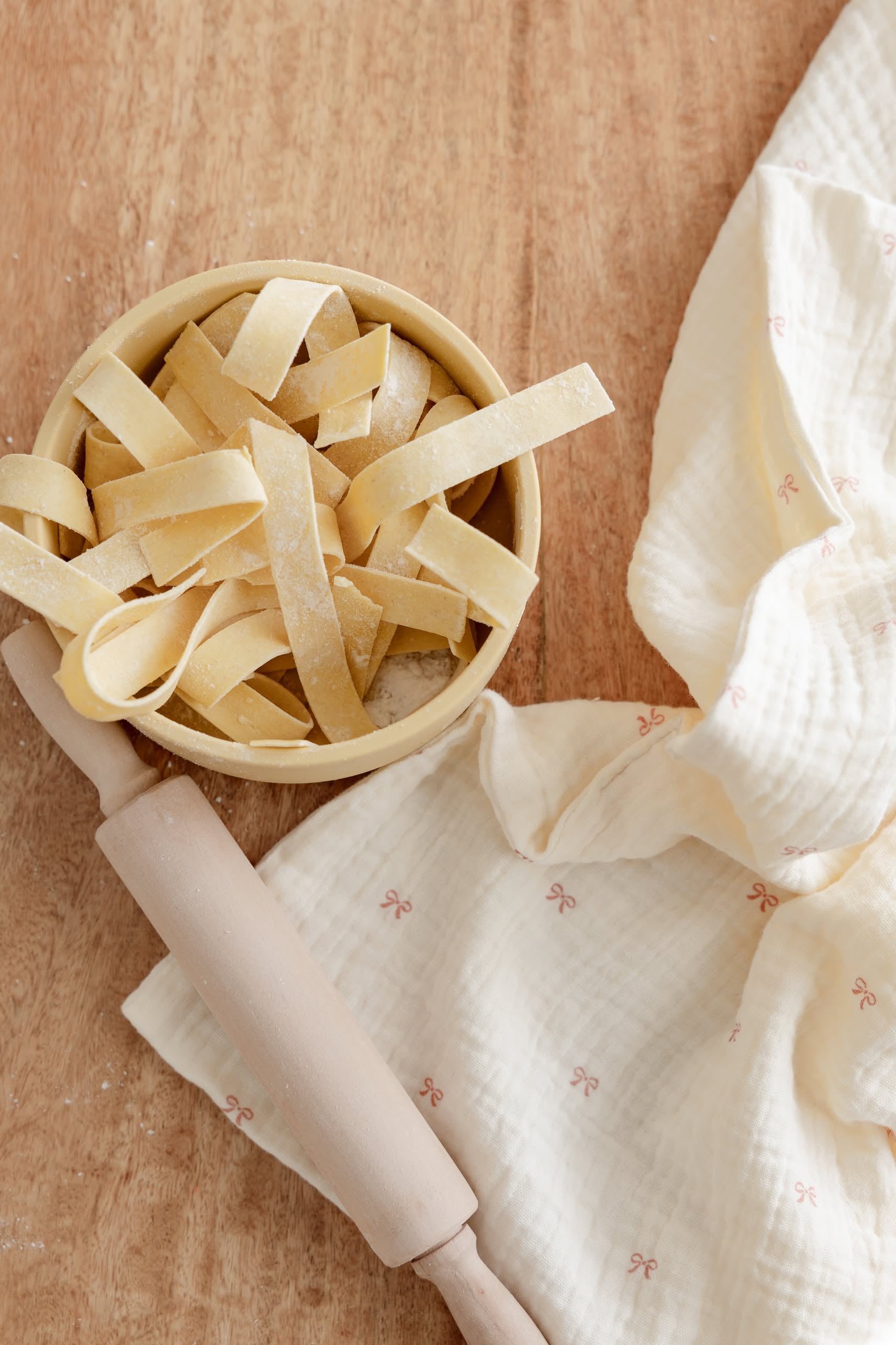 A bowl of uncooked tagliatelle sits on a wooden surface beside Soft Spot’s Soft Tea Towels (Set of 3) and a wooden rolling pin.