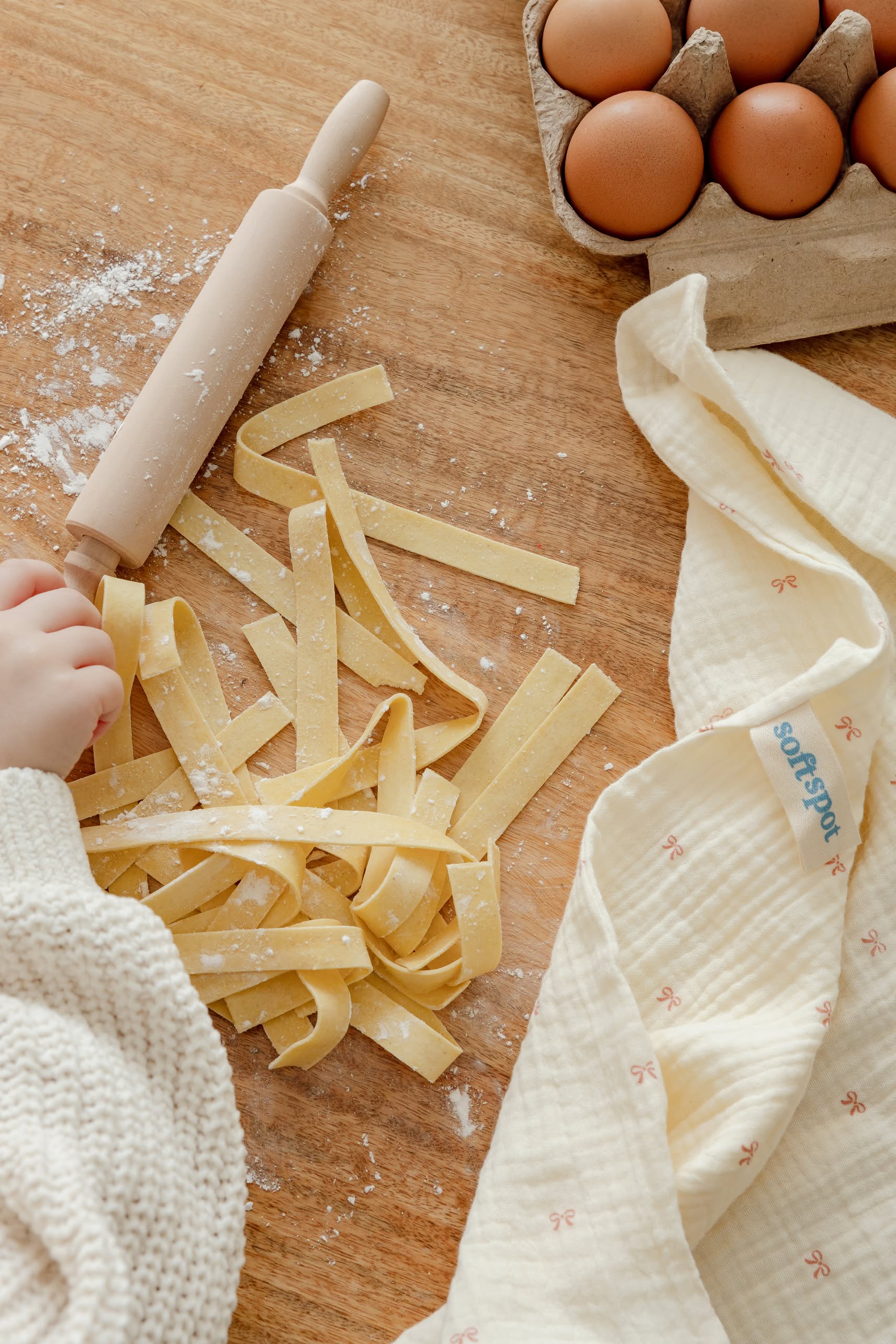 A childs hand near fresh pasta strips on floured wood, with a small rolling pin, eggs in a carton, and a Soft Spot Soft Tea Towel from the Soft Spot set of 3.
