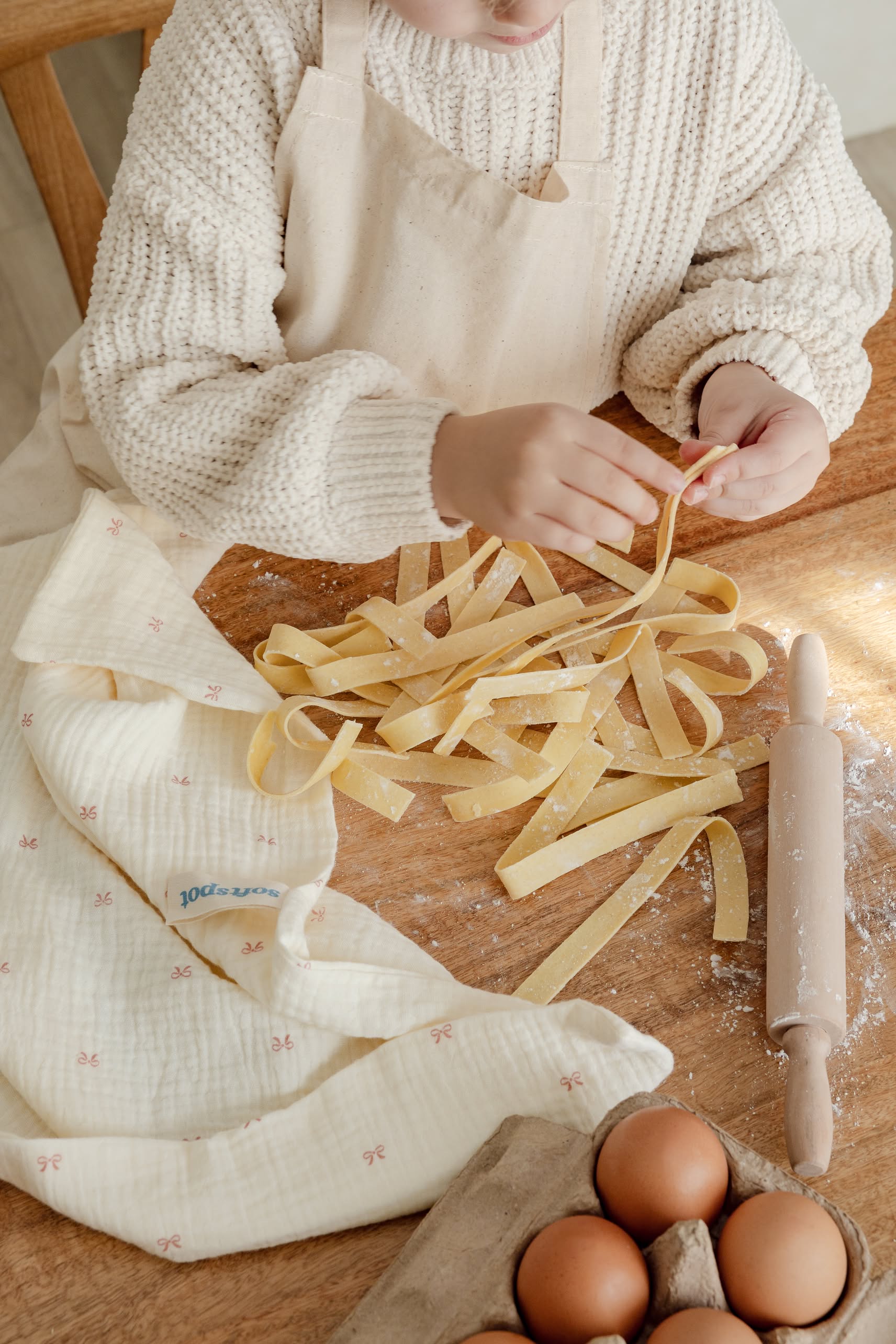 A person in a cream sweater and apron makes fresh pasta by hand on a wooden table, surrounded by a rolling pin, eggs, a cloth, and the Soft Spot Soft Tea Towels (Set of 3) from Soft Spot.