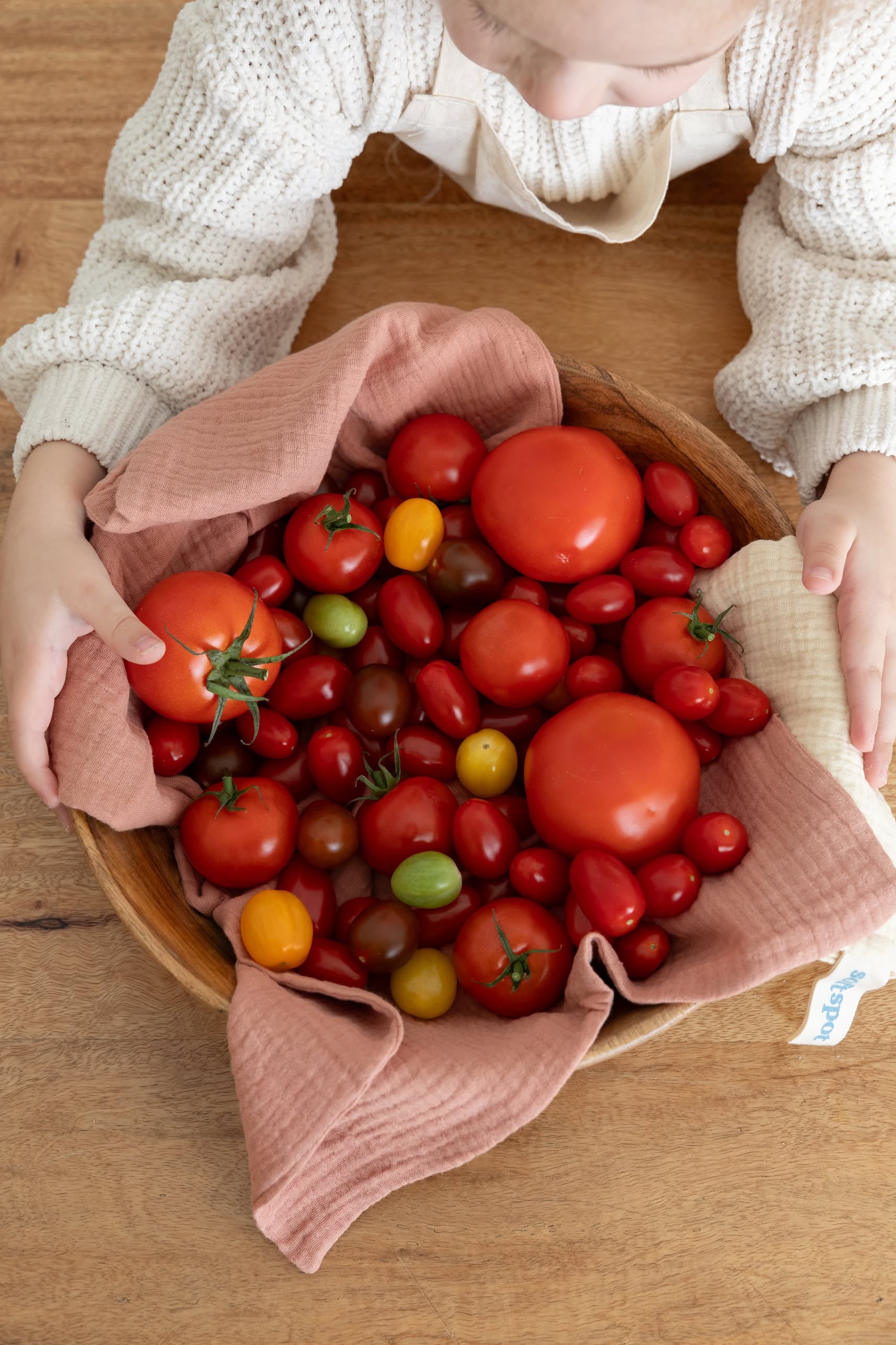 A child leans over a wooden bowl lined with a Soft Spot Soft Tea Towel, filled with assorted fresh tomatoes in red, yellow, and green, resting on a wooden table. The towel is part of the Soft Spot set of three.