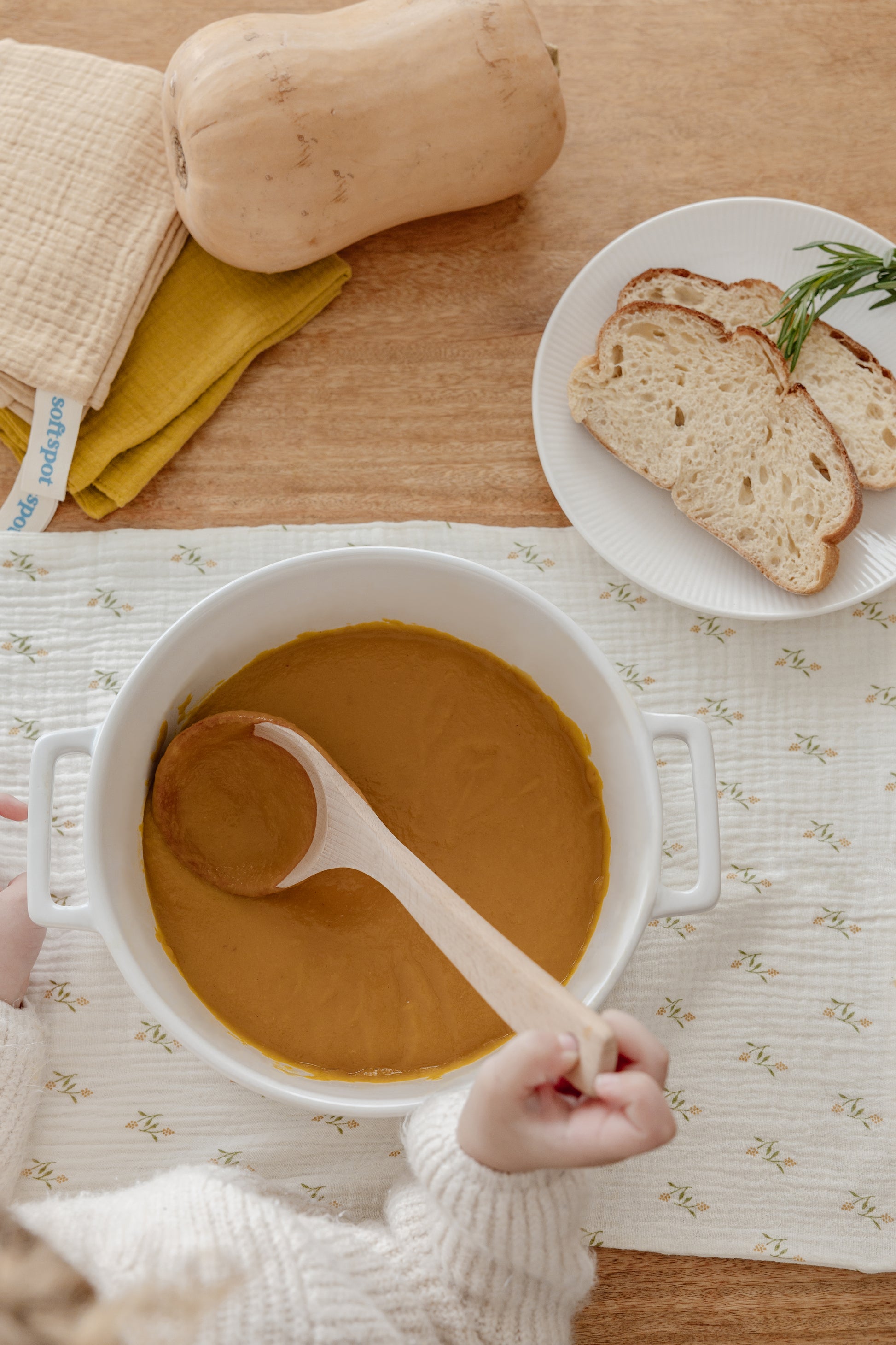 A loaf of bread rests on a white plate atop a folded Soft Spot Soft Tea Towel. Behind it are three onions in a bowl, a lidded white pot, and a wooden spoon, all displayed on a clean white surface. Set by Soft Spot.