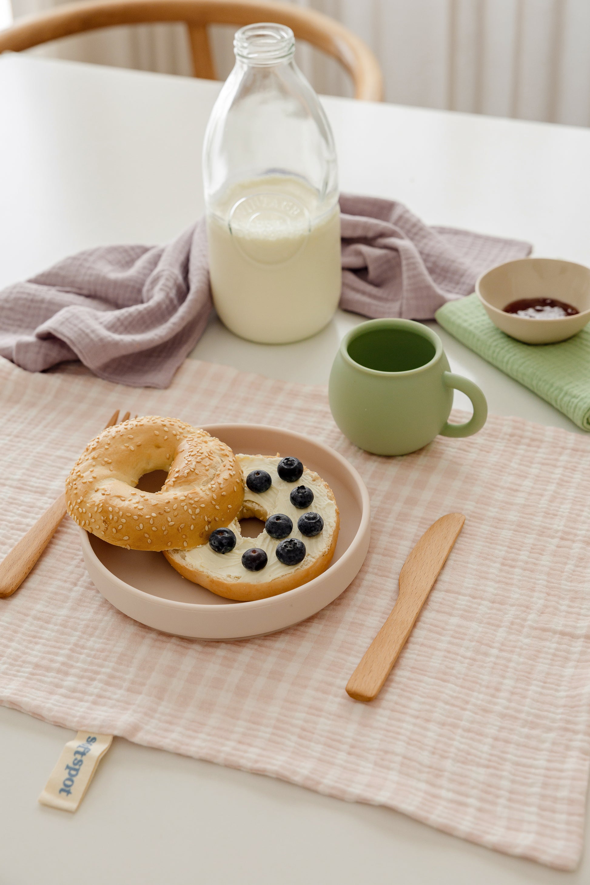 A sesame bagel half-topped with blueberries rests on a pink checkered placemat with wooden utensils, a green mug, a glass milk bottle, and a Soft Spot Soft Tea Towel (Set of 3) by Soft Spot on a white table.