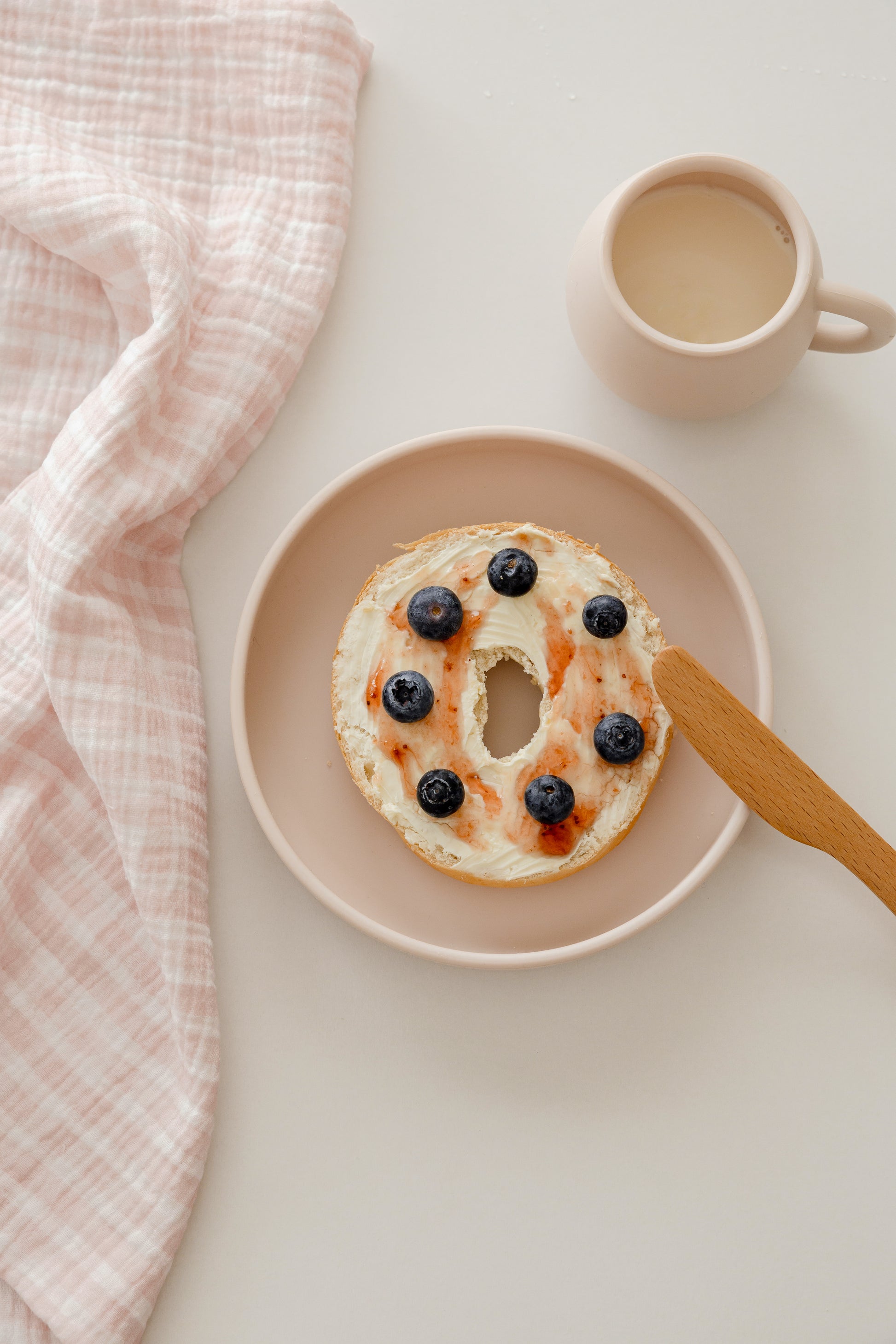 A bagel with cream cheese, blueberry jam, and fresh blueberries sits on a pink plate with a wooden butter knife. A pink mug of coffee and a Soft Spot Soft Tea Towel (Set of 3) from Soft Spot are nearby on a light surface.