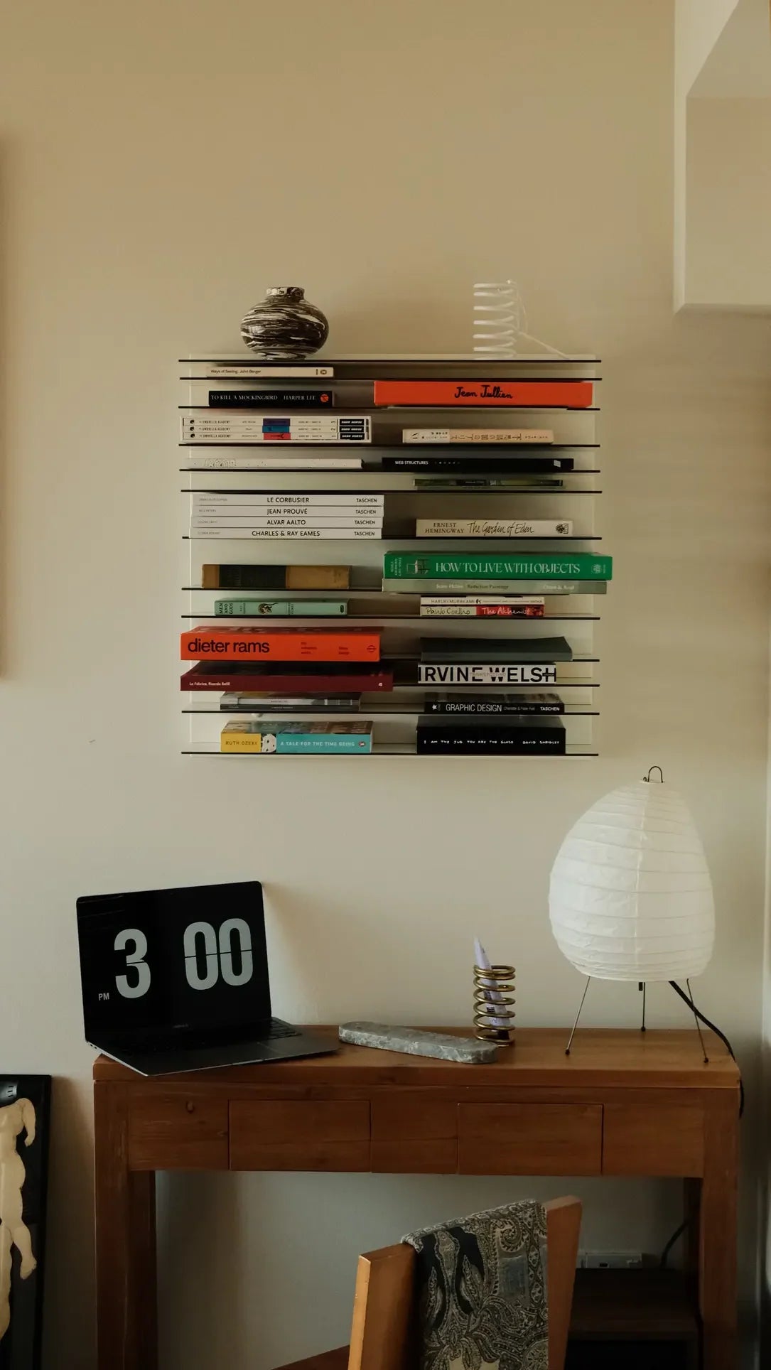 A wooden desk with a lamp, a digital clock showing 3:00, and a notepad sits beneath the Spectrum Paperback Wallsystem 60x60 by Spectrum, which holds neatly stacked books and small decorative items in a cozy room.