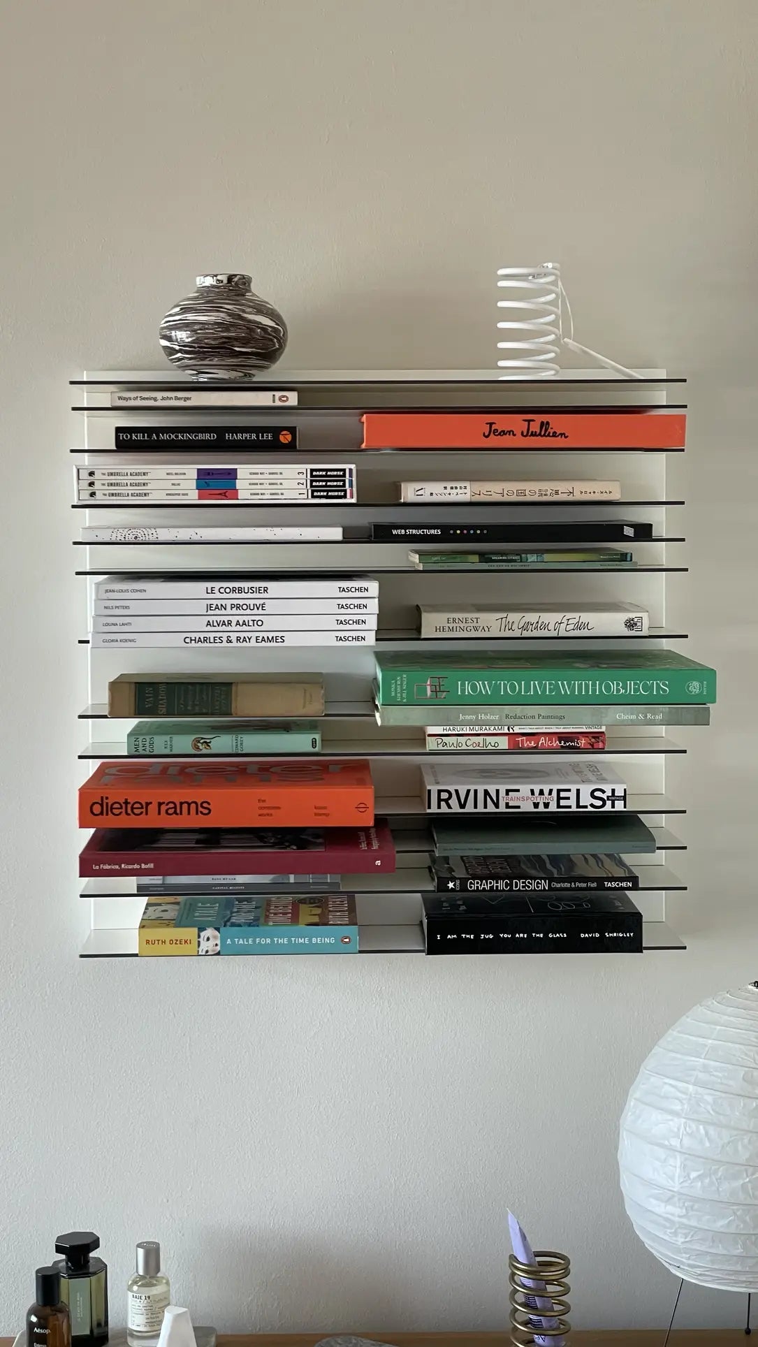 The Spectrum Paperback Wallsystem 60x60 by Spectrum showcases art, design, and architecture books in horizontal stacks. A striped vase and white coiled sculpture rest above, with a round lamp at the bottom right to enhance the minimalist aesthetic.