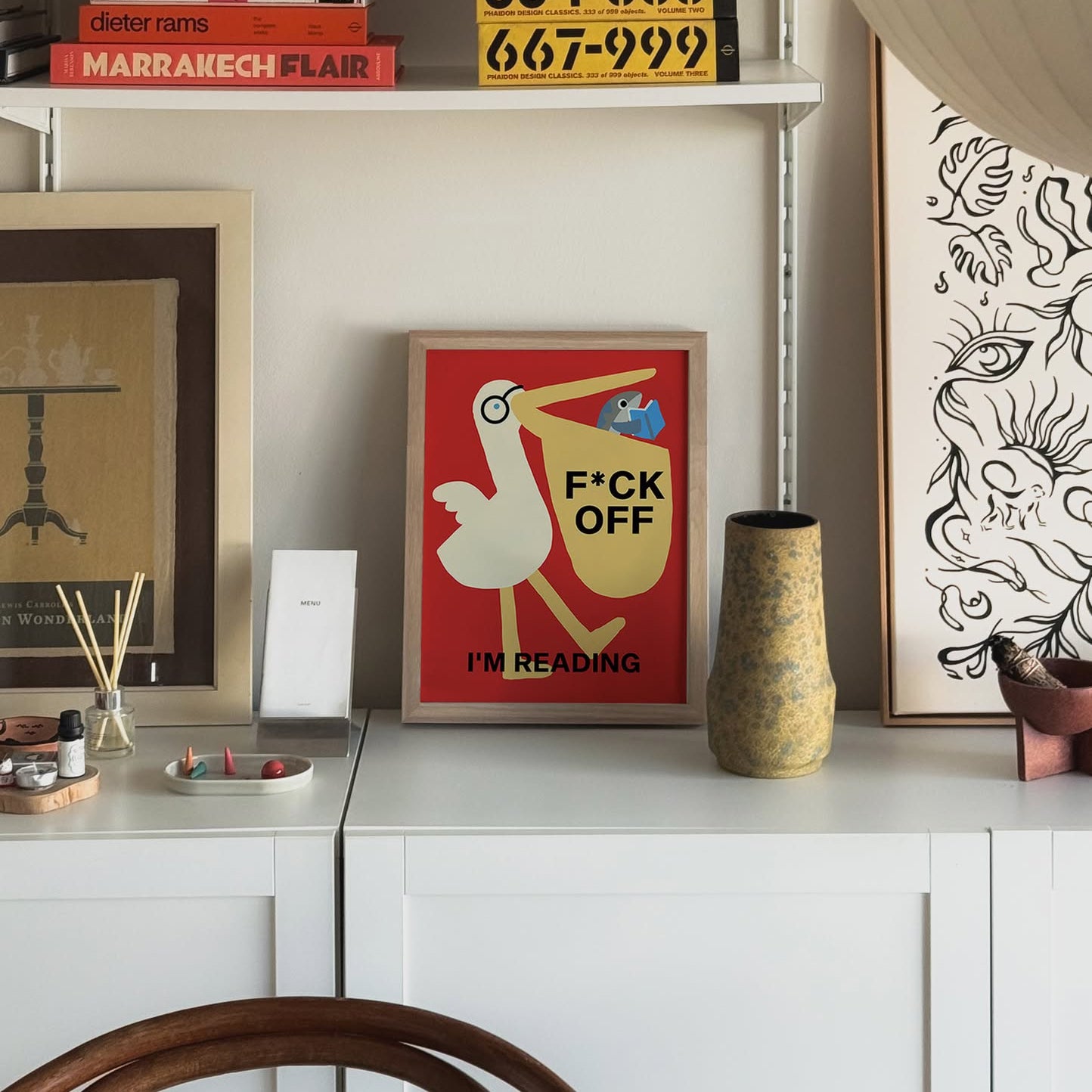 The Stacked Store Fuck Off Im Reading - Pelican Potter Poster (30x40) is displayed on a white cabinet, surrounded by books, art, and decor in a stylish room. The print features a book-loving bird holding a book that says FUCK OFF, IM READING.