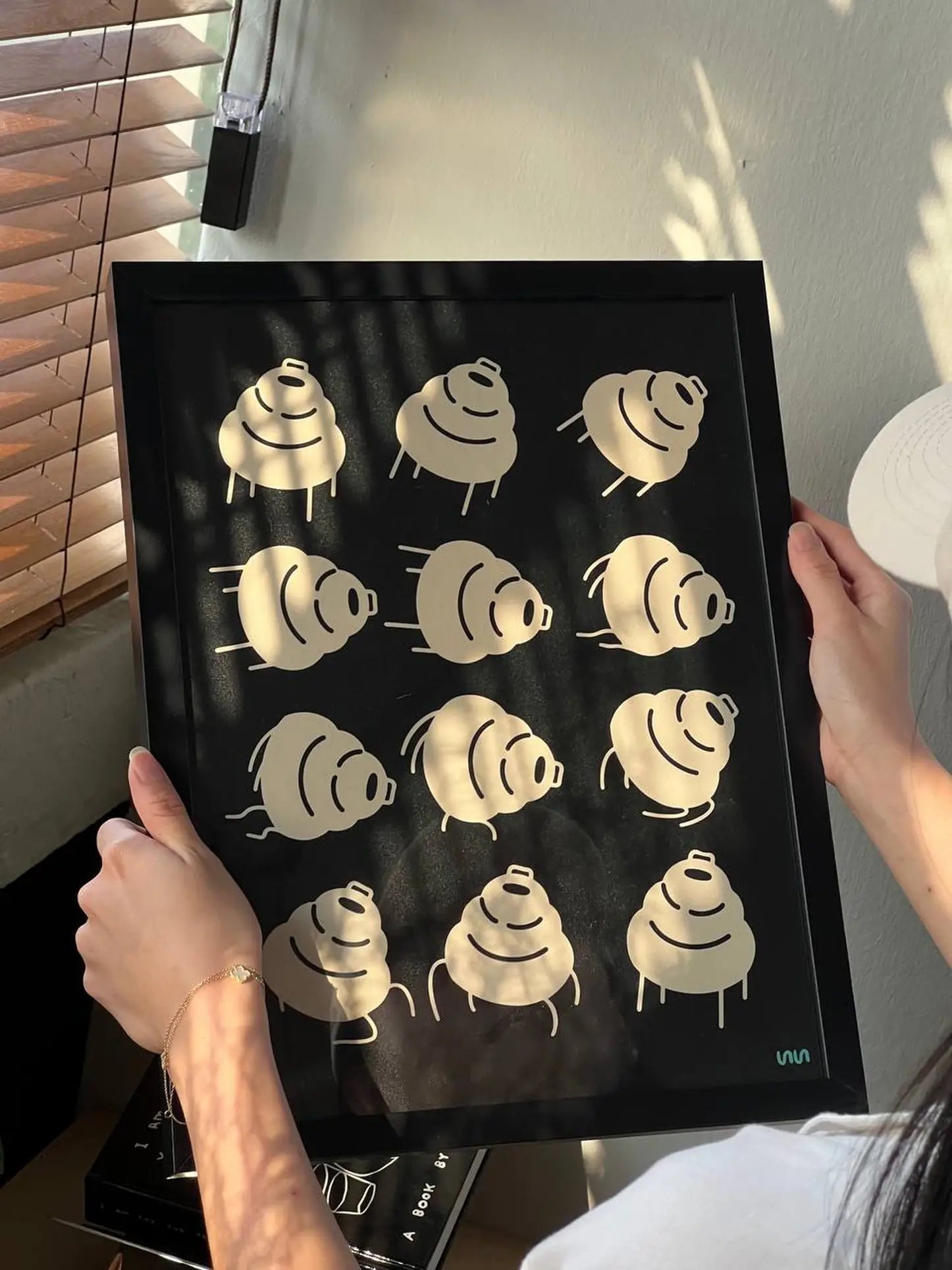 A person holds the Stacked Store Trip Poster by Stacked Store, showcasing a grid of twelve stylized beige croissants on a black background; sunlight from blinds casts minimalist shadows, enhancing its modern appeal.