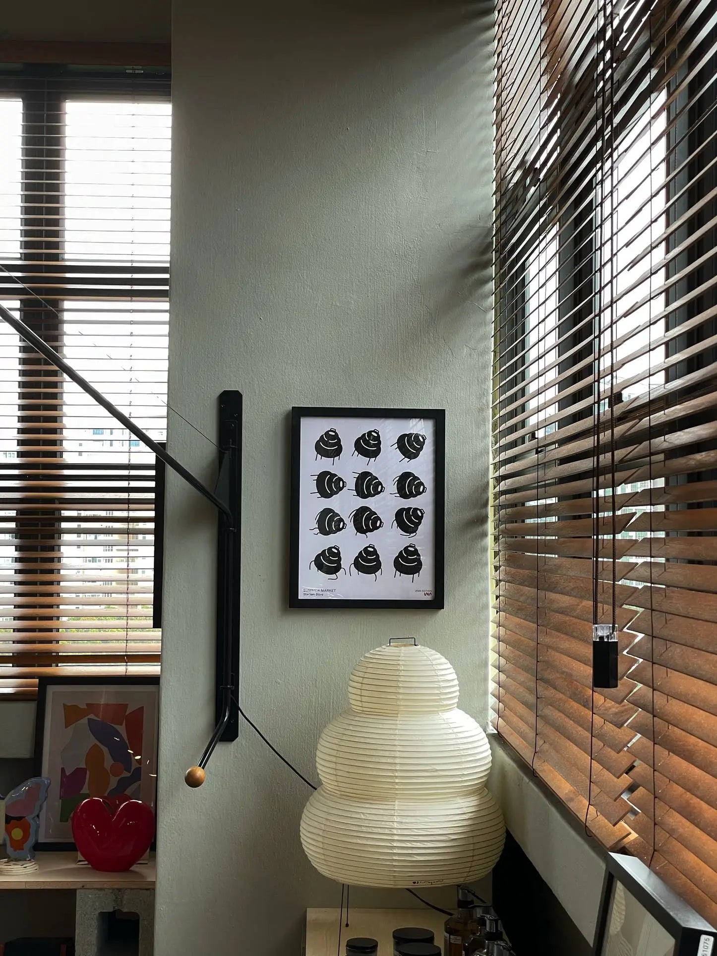 A cozy room corner with wooden blinds, a Stacked Store Summer Market 2024 Edition Poster on the wall, a minimalist Akari 24N lamp on a shelf, and art objects like a red heart sculpture. Daylight softly filters through the window.