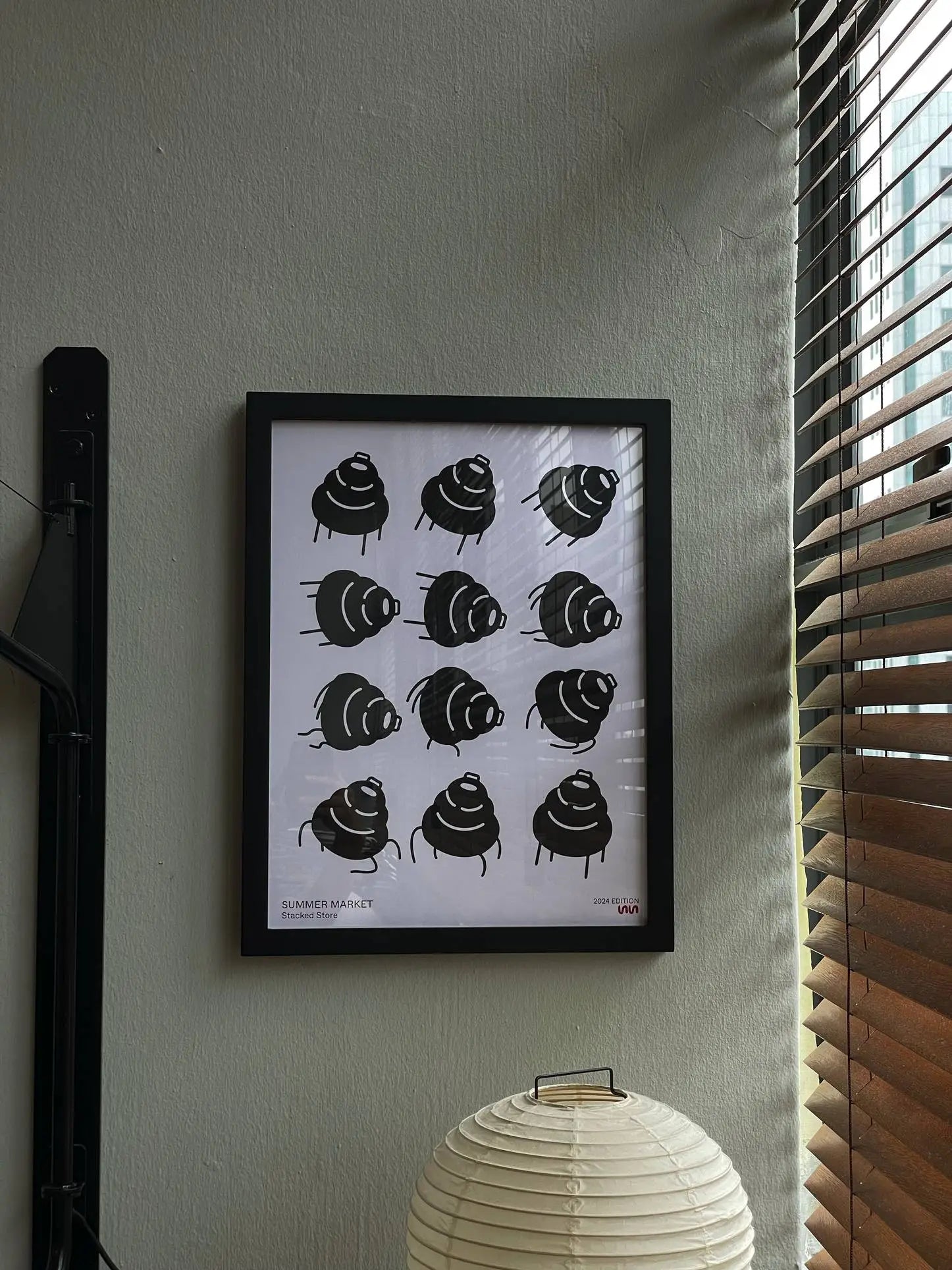A framed Stacked Store Summer Market 2024 Edition Poster with 12 dark-hued shells in a 3x4 grid hangs on a gray wall by a window with wooden blinds, complemented below by an Akari 24N lamp for minimalist style.