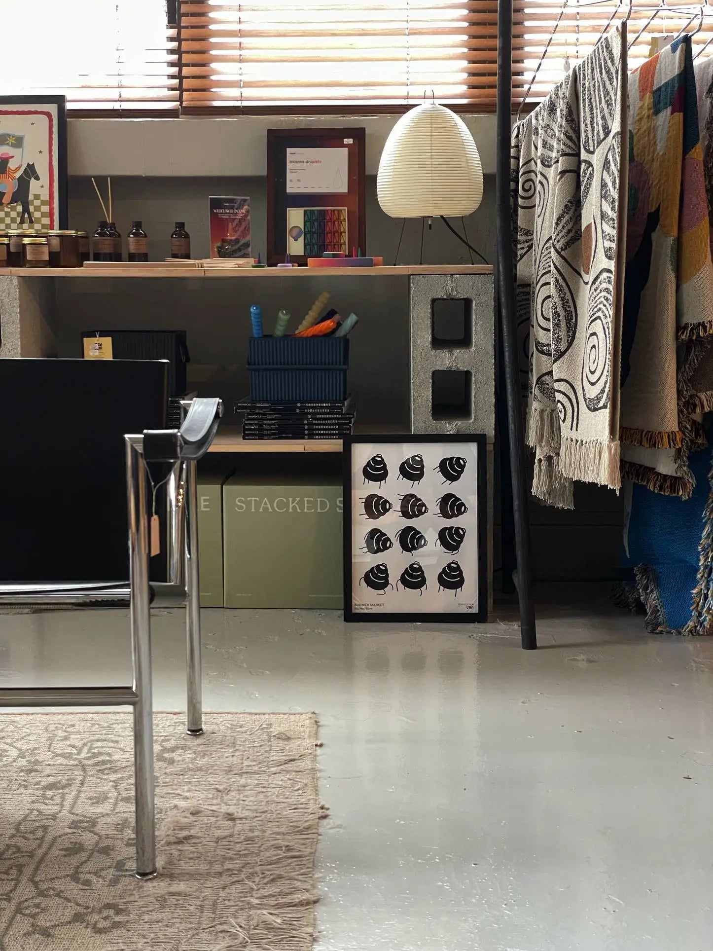 A modern room with minimalist style has a metal chair, a shelf with books, bottles, and an Akari 24N lamp. Patterned blankets hang nearby; the Stacked Store Summer Market 2024 Edition Poster leans against the wall beside framed art.