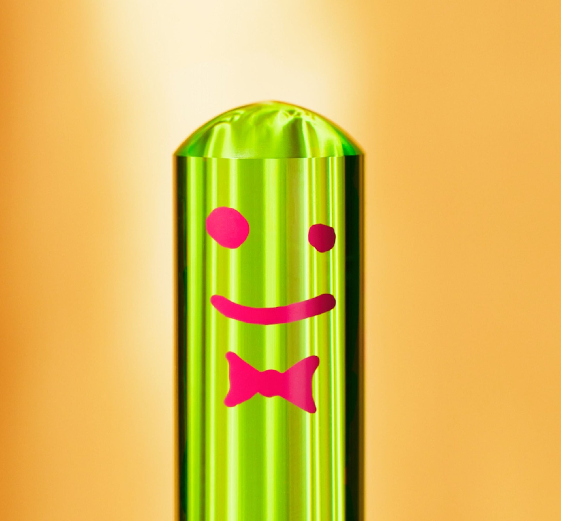 The Staff The Toilet Brush - Green by Staff is a shiny green cylindrical brush with a pink smiley face, bow tie, and an acrylic handle, displayed against an orange background.
