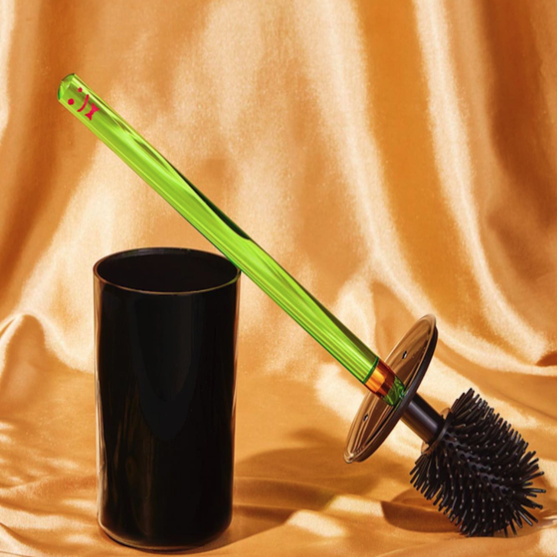 The Staff The Toilet Brush - Green by Staff features a replaceable brush head and neon green acrylic handle, shown with its black cylindrical holder on a shiny gold fabric background.