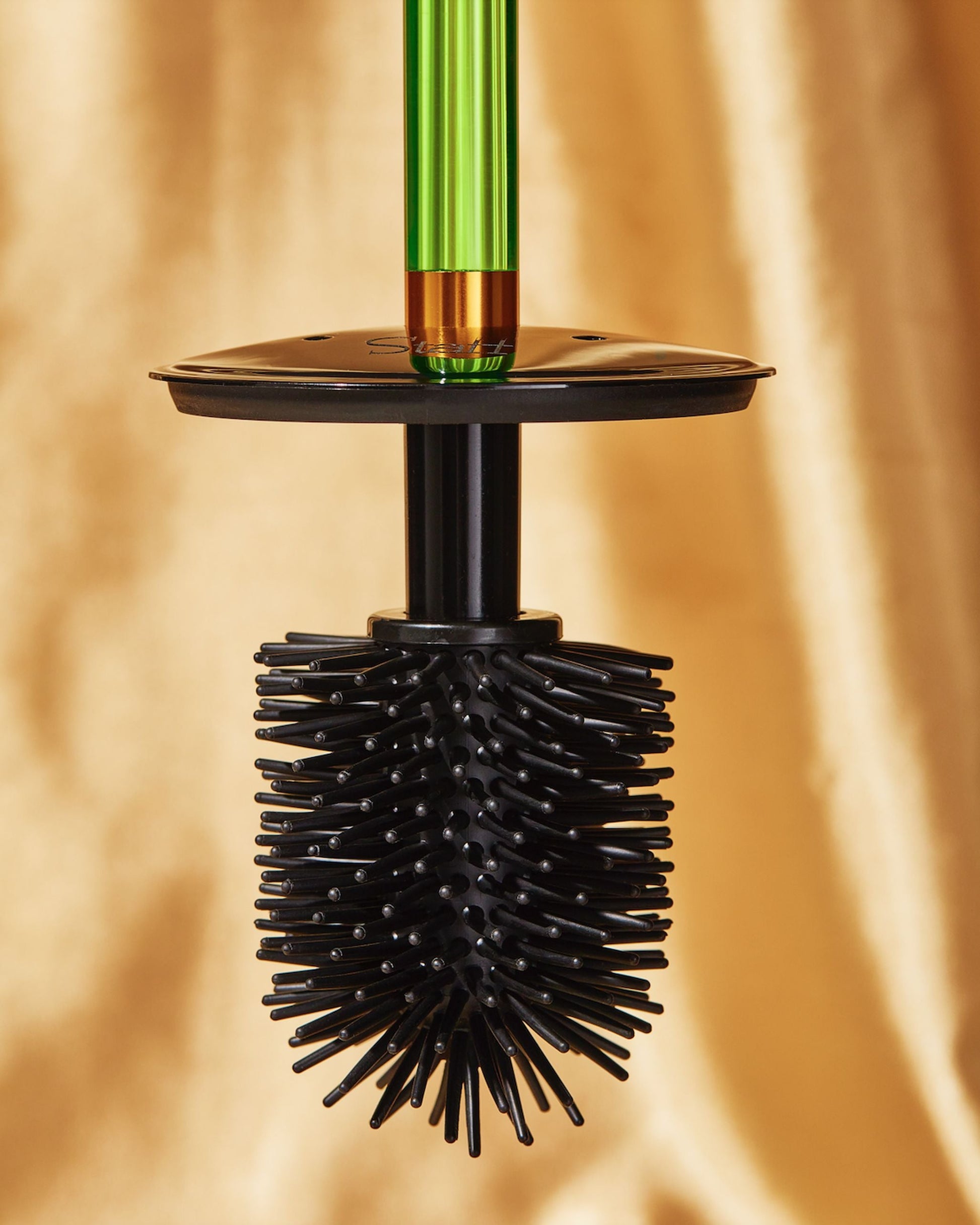 A close-up of Staff The Toilet Brush - Green by Staff, featuring flexible bristles, a green acrylic handle, and a replaceable brush head, set against a gold fabric background.
